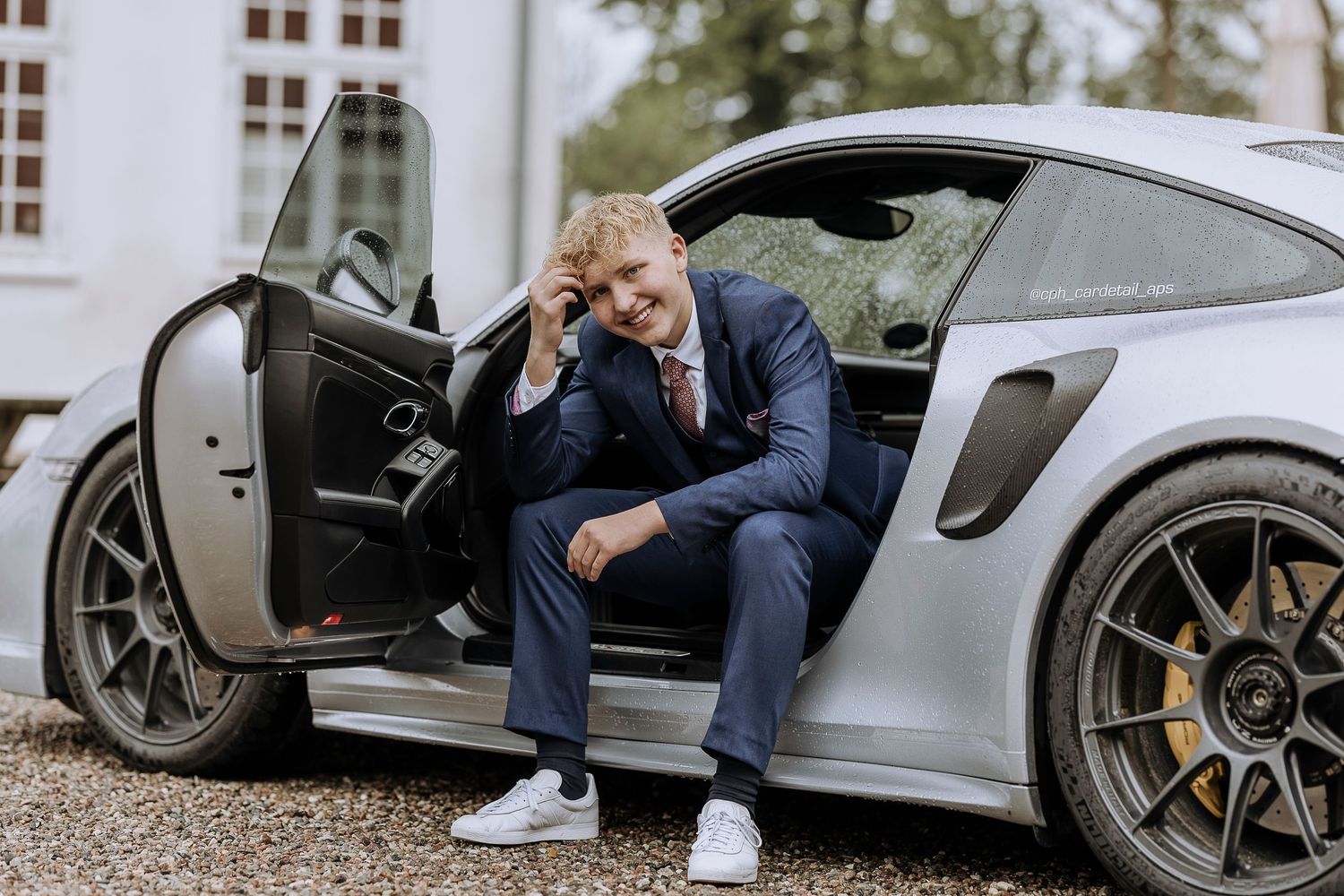 Person in blue suit sits in gray sports car with door open, showing contemplative pose at building exterior.