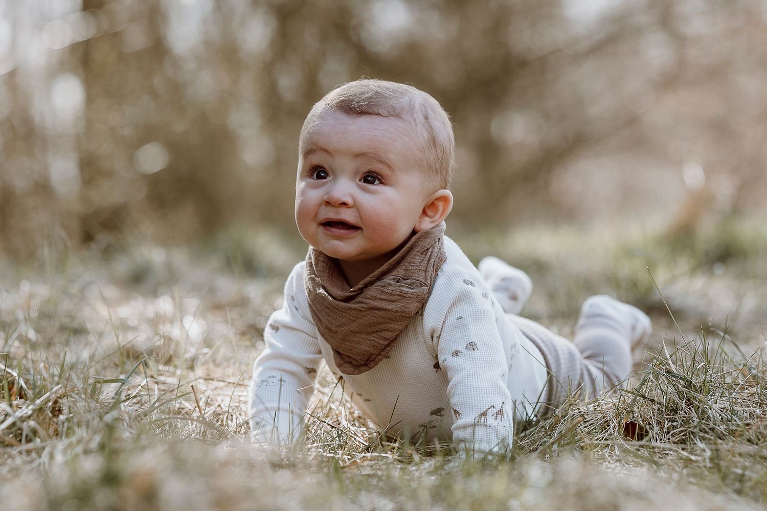 Baby wearing white outfit lies on grass while looking up with a joyful expression during an outdoor photo session.