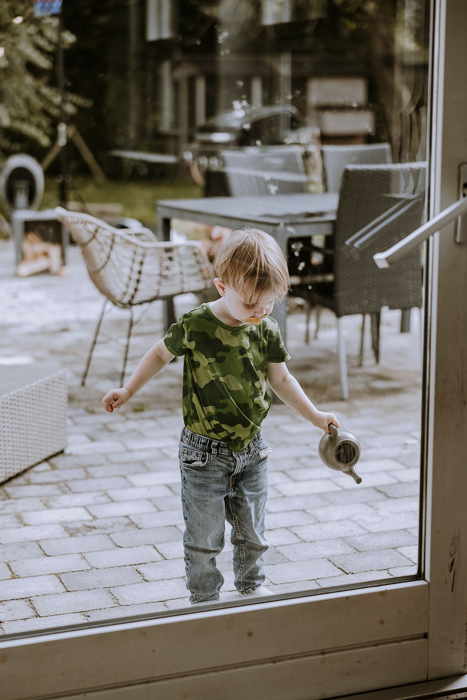 Young child in camouflage shirt playing on outdoor patio through window view.