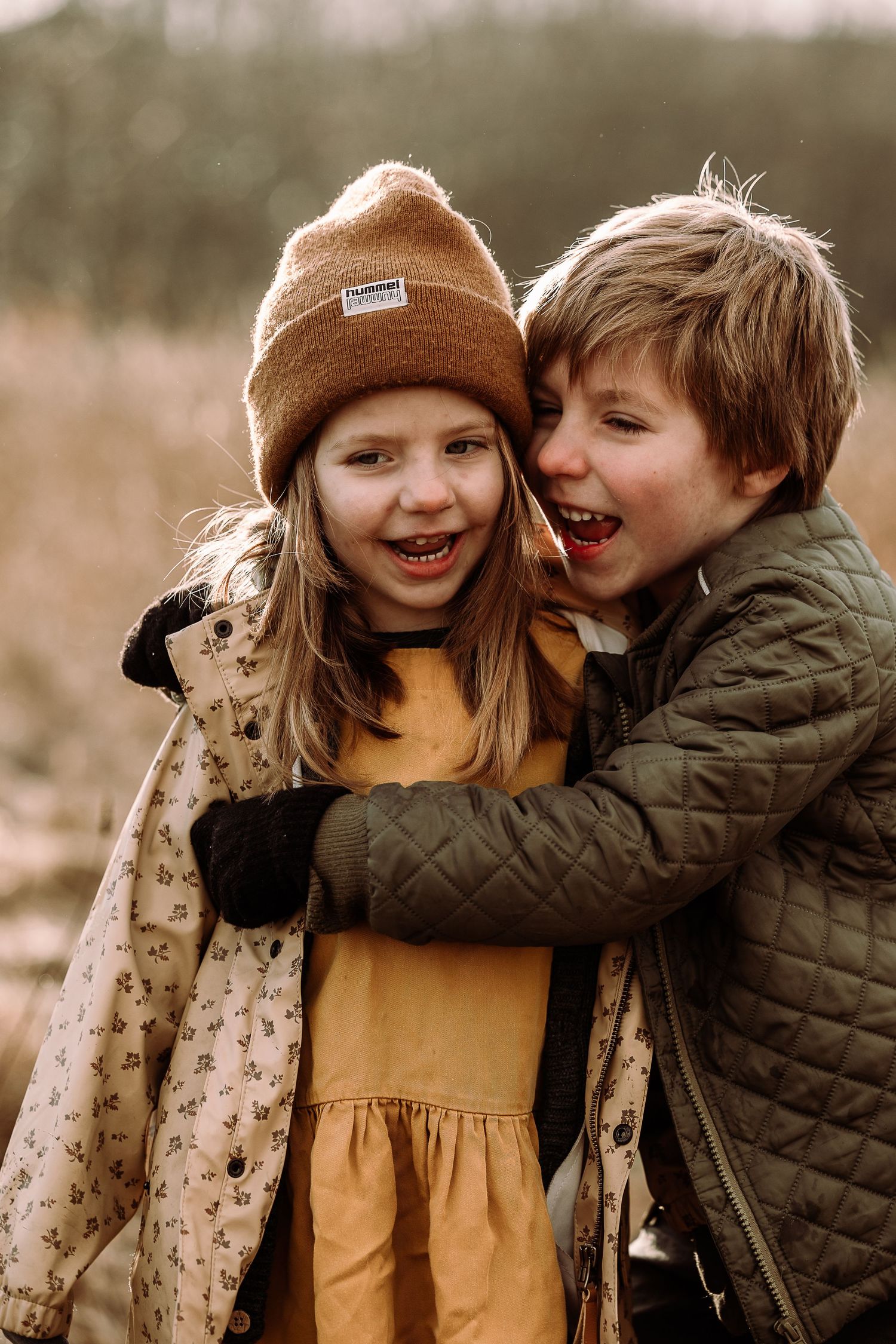 Two children share a playful moment outdoors in autumn wearing a brown beanie and quilted jacket.