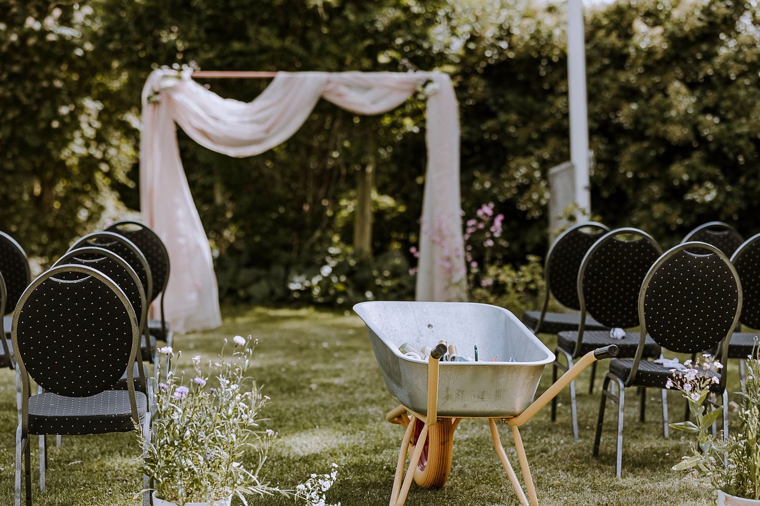 Outdoor wedding ceremony setup with white draped arch and black chairs on a lawn.