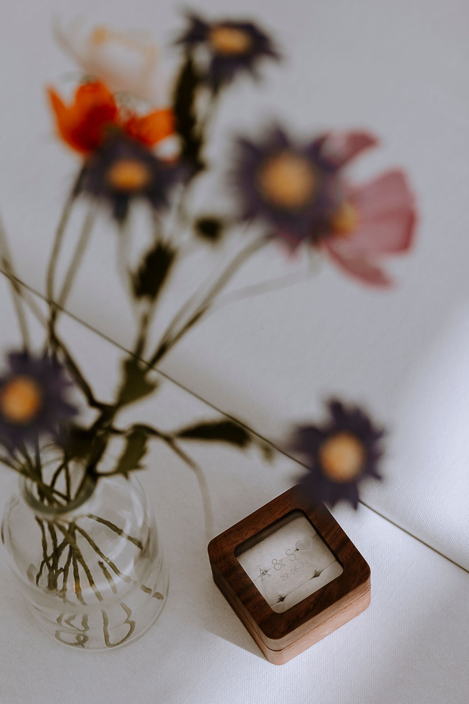 Delicate wildflowers in a clear glass vase next to a wooden ring box on a white surface.