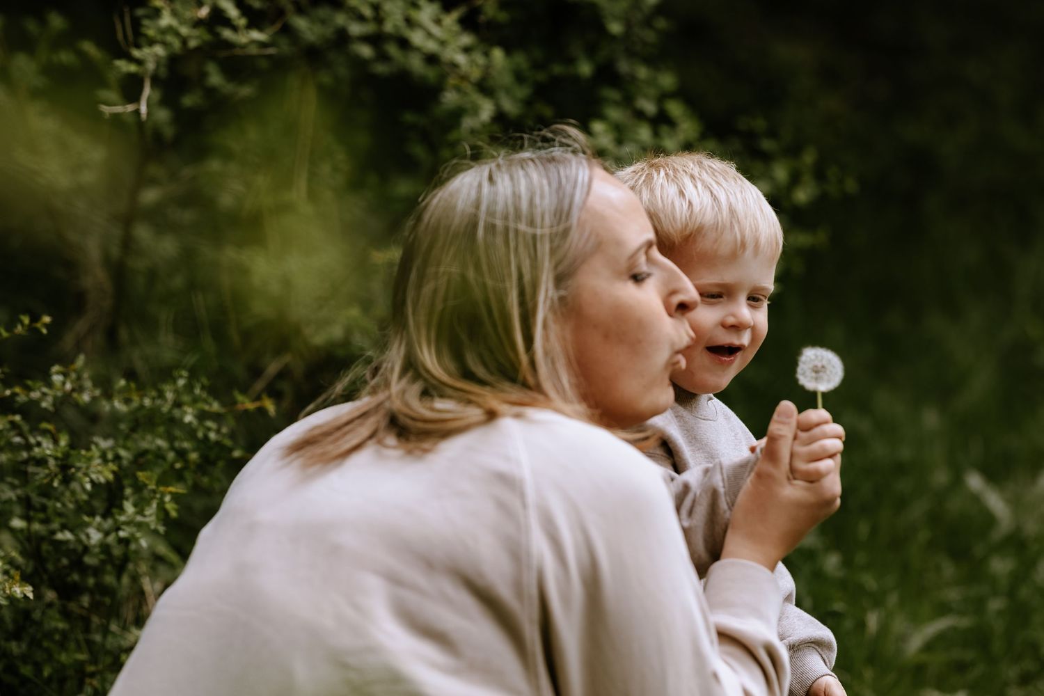 Two people blowing dandelions together in a magical outdoor moment surrounded by greenery.