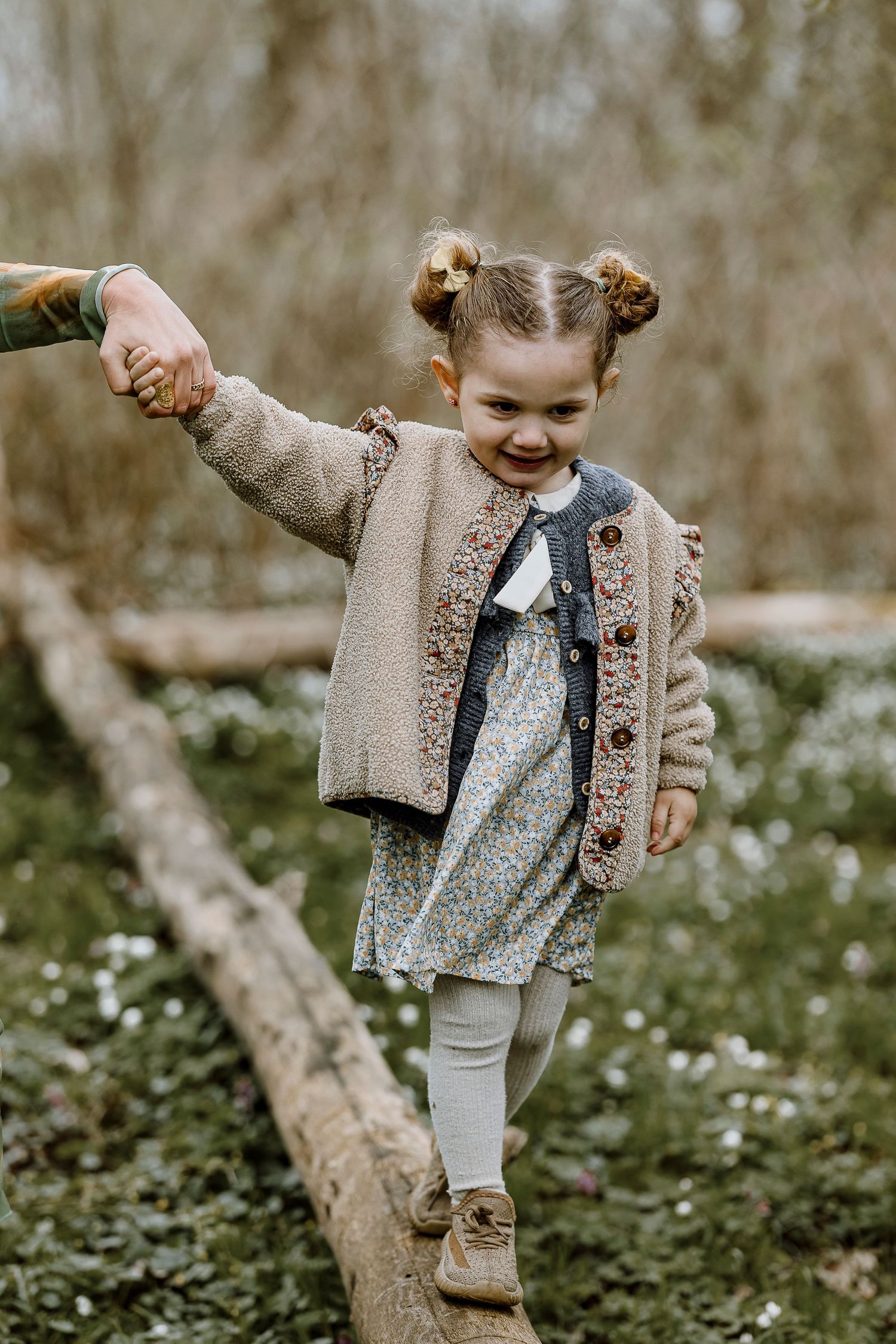 A child in a beige cardigan and floral dress plays joyfully among blooming wildflowers in a natural outdoor setting.