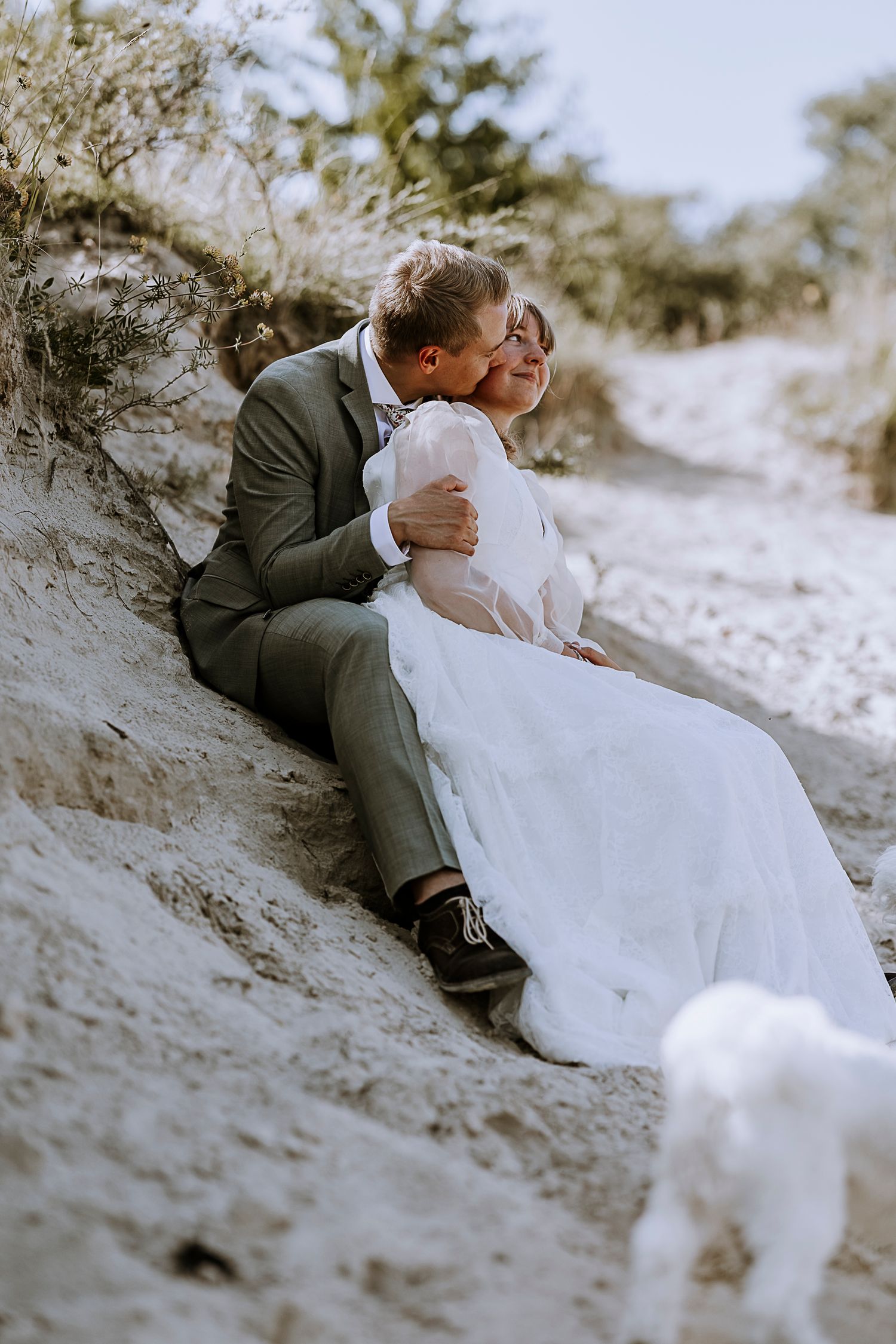 Couple in wedding attire sitting on a sandy slope with a white dog in the background.