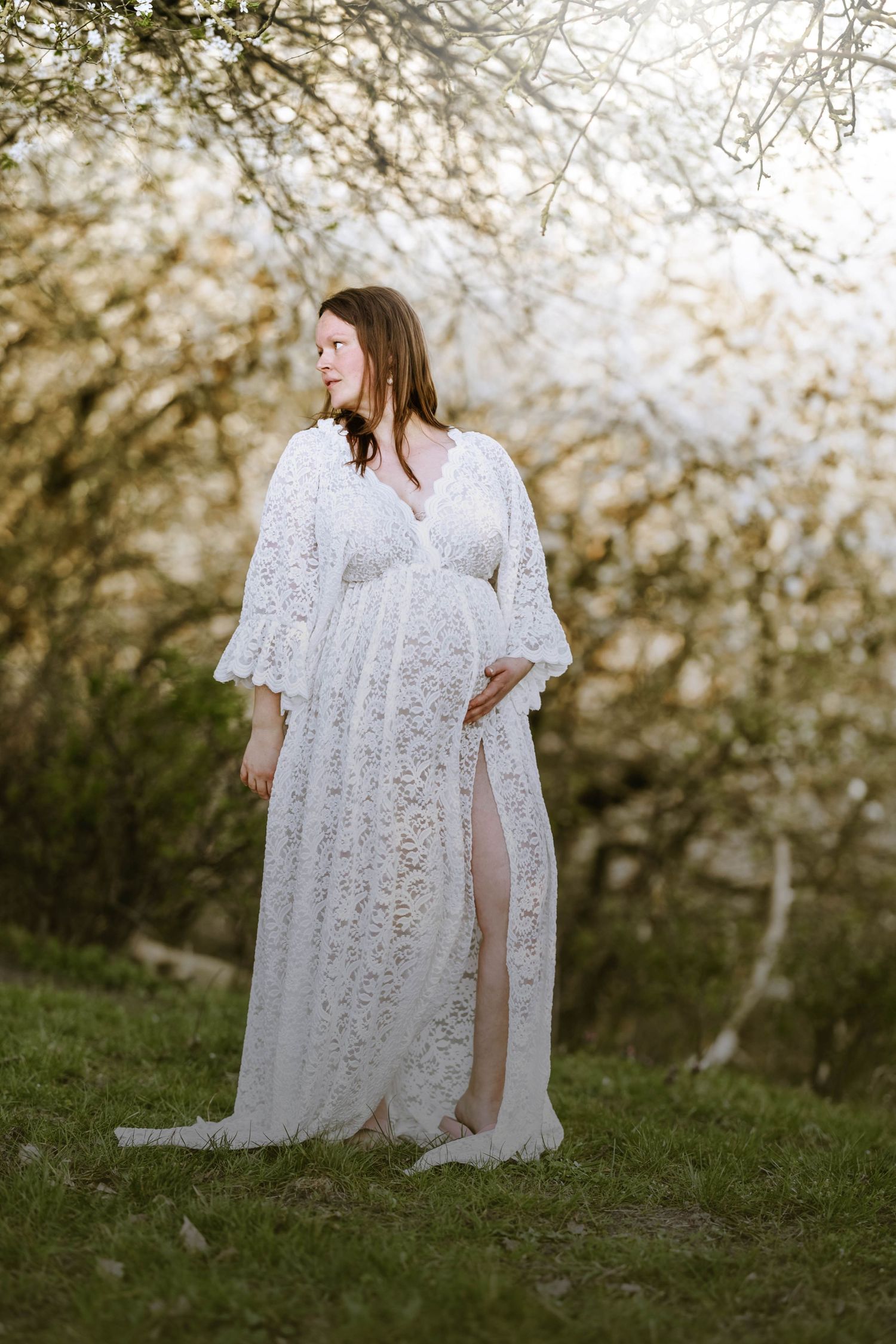 Woman in flowing white lace maternity gown stands gracefully on grassy hillside.