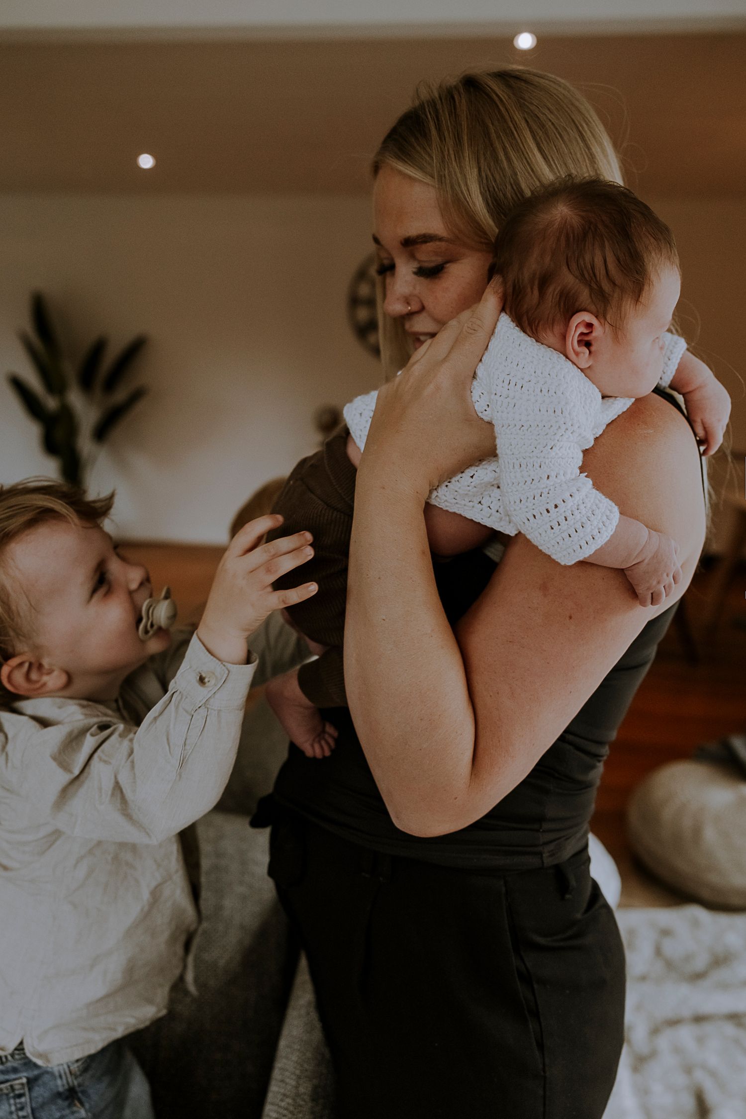 Mother in black top holds baby in white sweater while toddler reaches up playfully.
