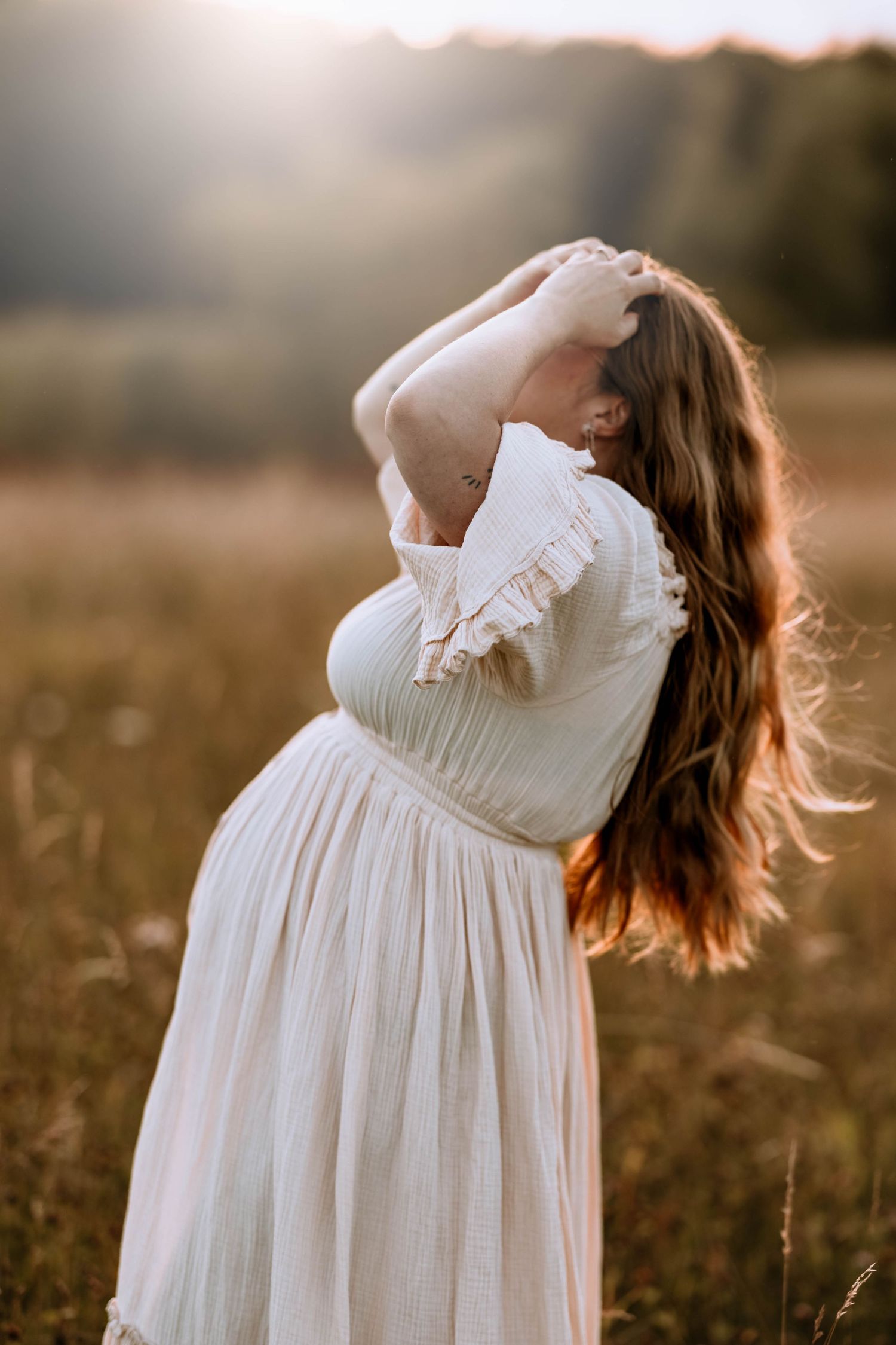 Pregnant woman in white dress arching back in golden sunlit field.