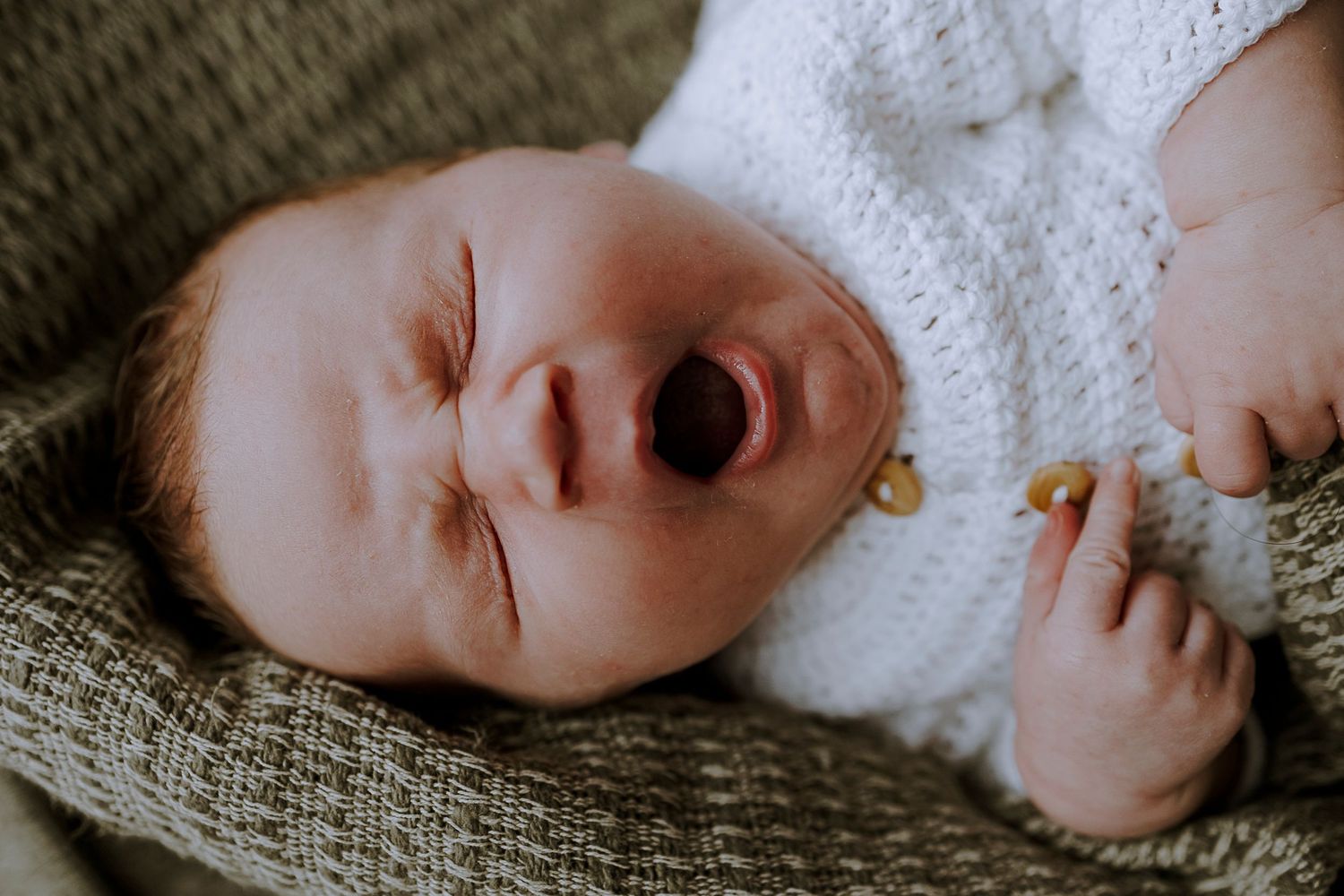 Close-up of yawning newborn baby wearing white knitted clothing on textured blanket.