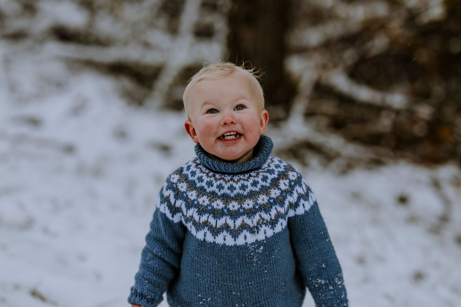 Toddler in blue Fair Isle sweater smiling outdoors in winter snow.