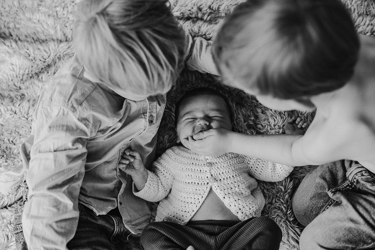 Black and white portrait of siblings bonding with baby on carpet.
