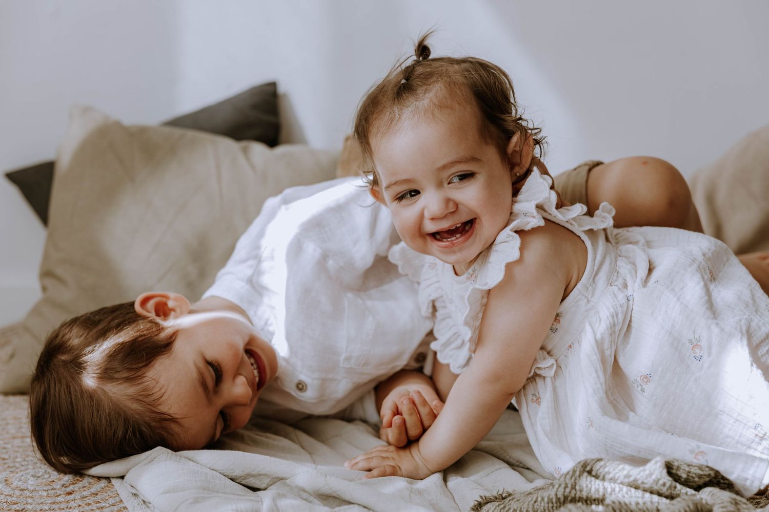 Two children in white clothing lying on bedding while sharing a sweet candid moment together.