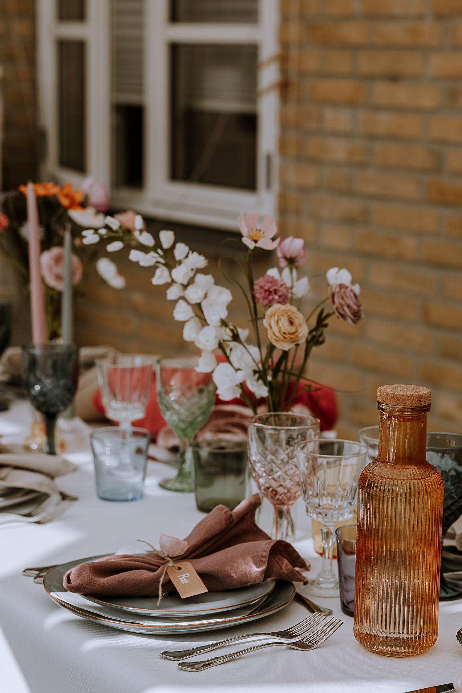 Elegant table setting with amber bottle, crystal glasses and floral arrangements against brick wall.