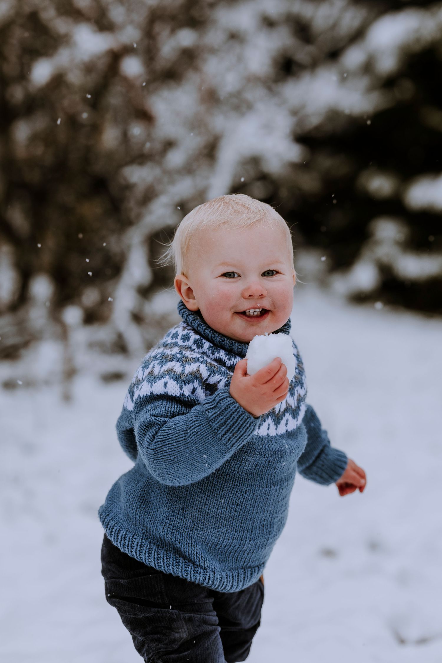 Small child wearing blue Fair Isle sweater holds snow while playing outdoors in winter wonderland.