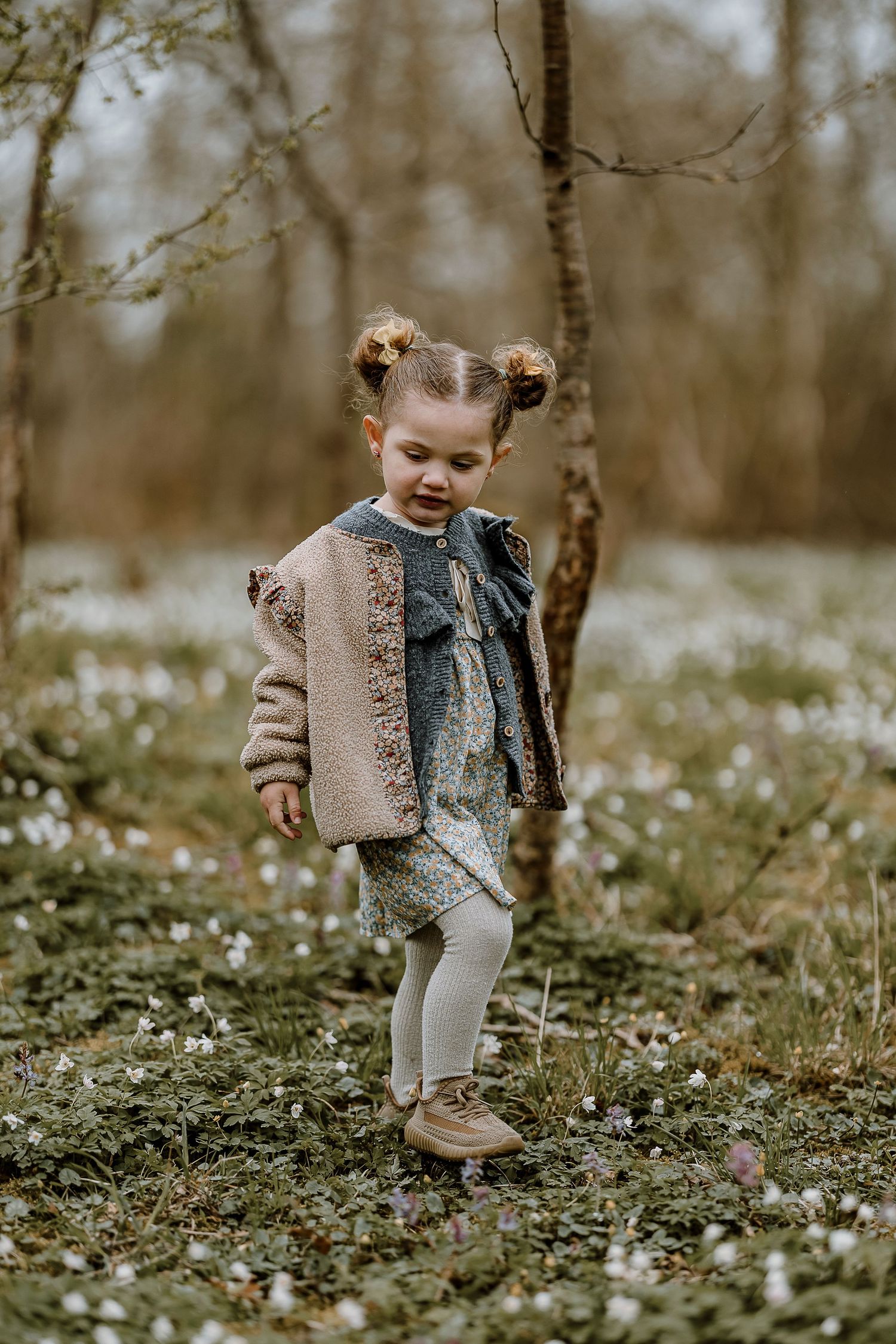 A child in a beige sweater and boots walks through a field of small white wildflowers on a misty spring day.