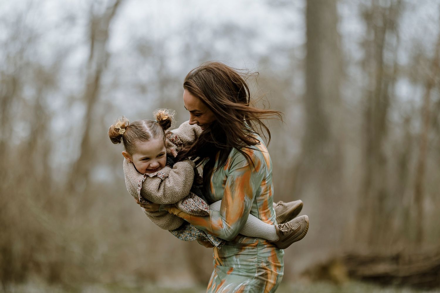 A figure in a floral dress holds and plays with a small dog while standing in a wooded area on an autumn day.