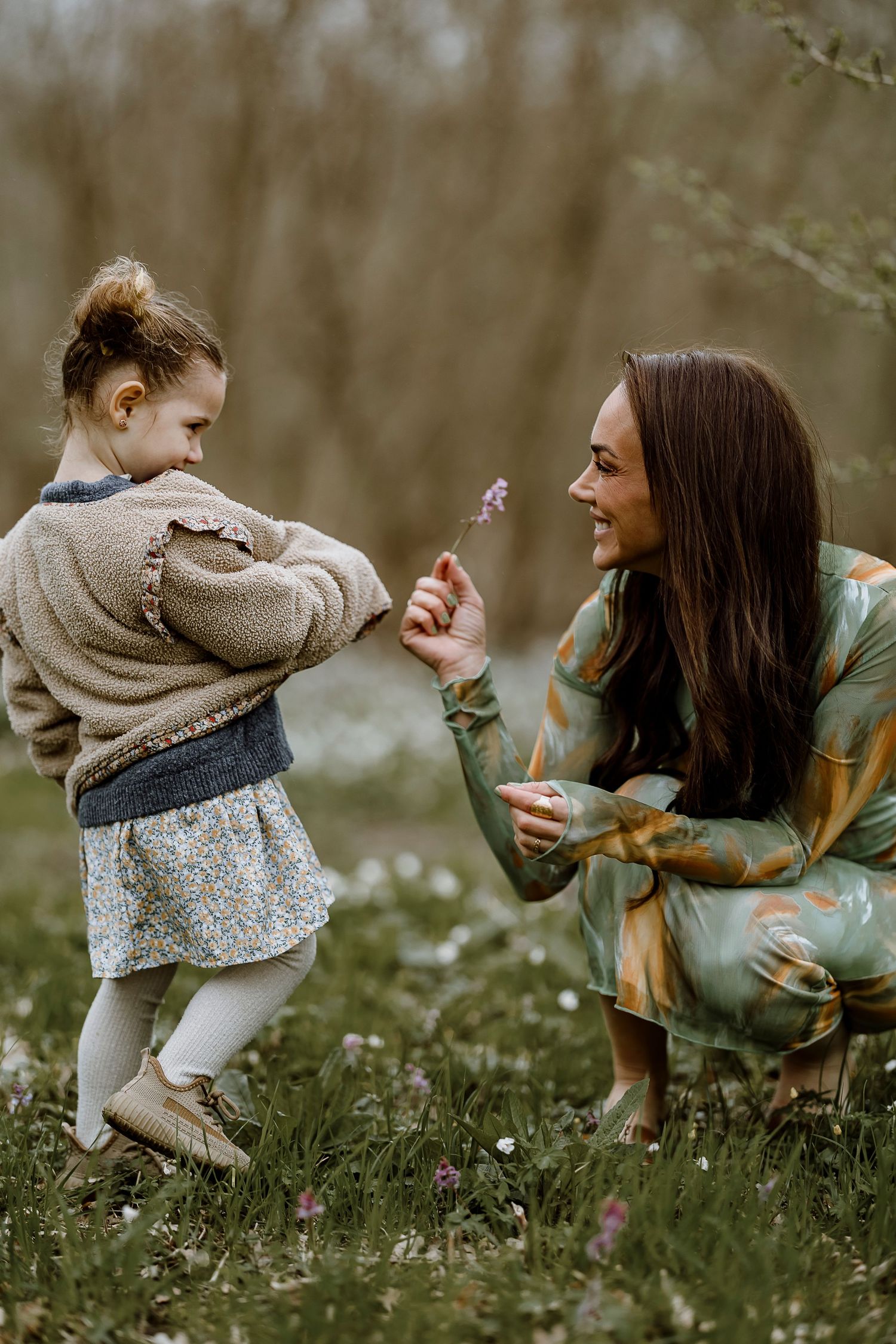 A woman and child sit in a grassy meadow blowing dandelions together on a spring afternoon.