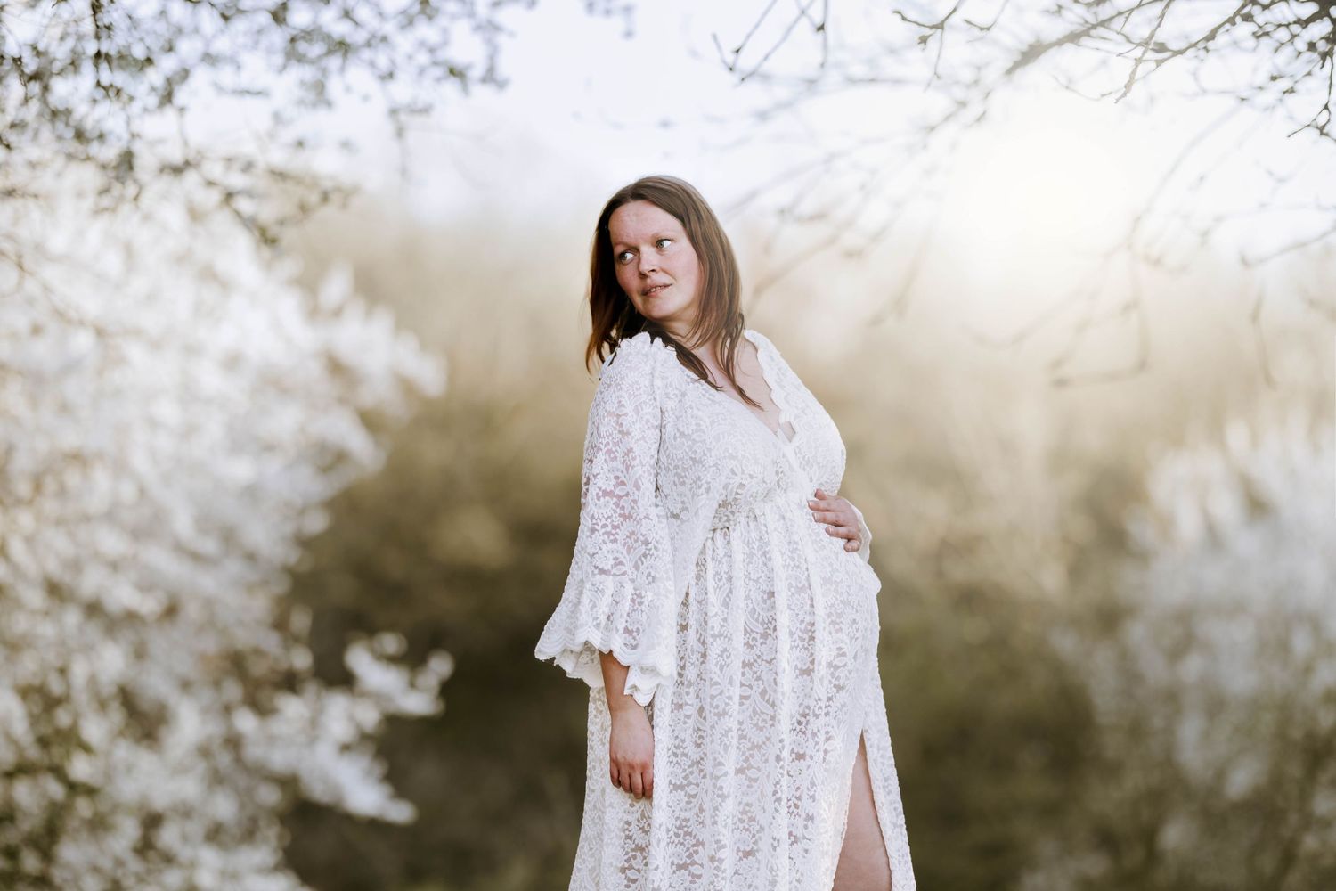 Maternity portrait of woman in white lace dress against spring blossoms.