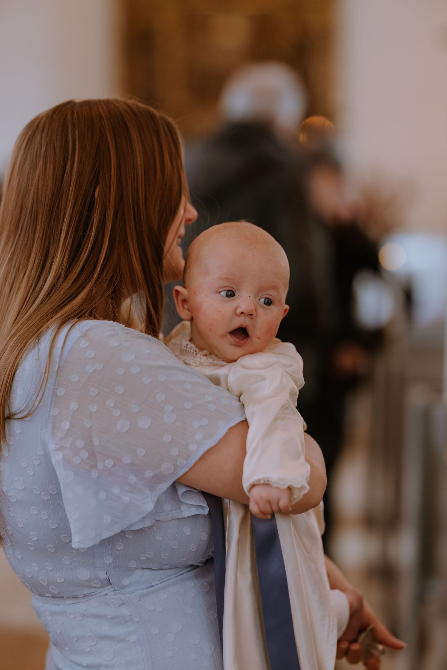 A woman in a blue dress holds a wide-eyed baby in a white christening gown at an indoor ceremony.
