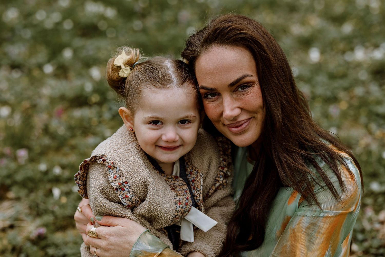 A parent and child sit together outdoors surrounded by greenery while holding a gray cat.