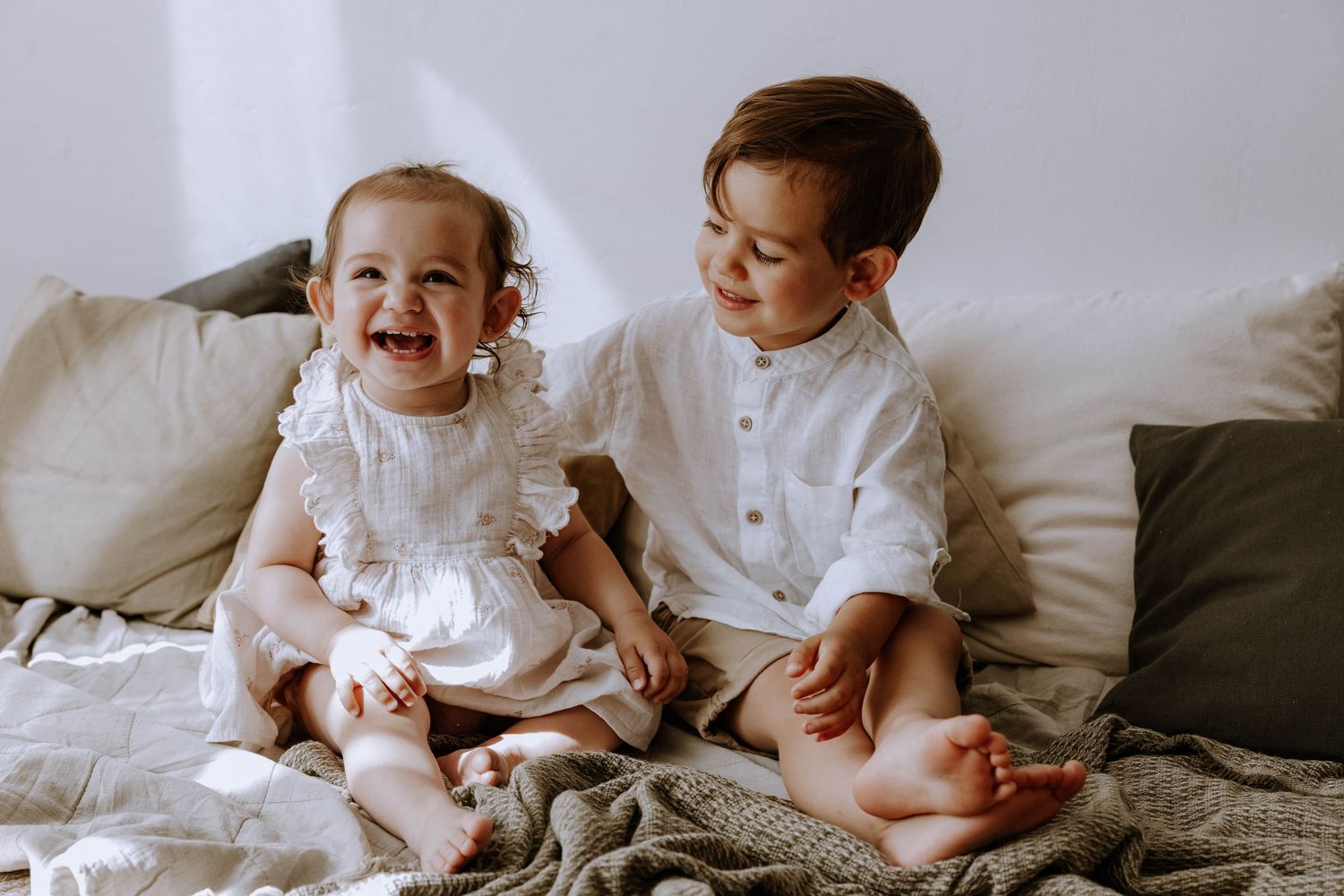 Two young children in white outfits sit together on a bed with neutral bedding and pillows.
