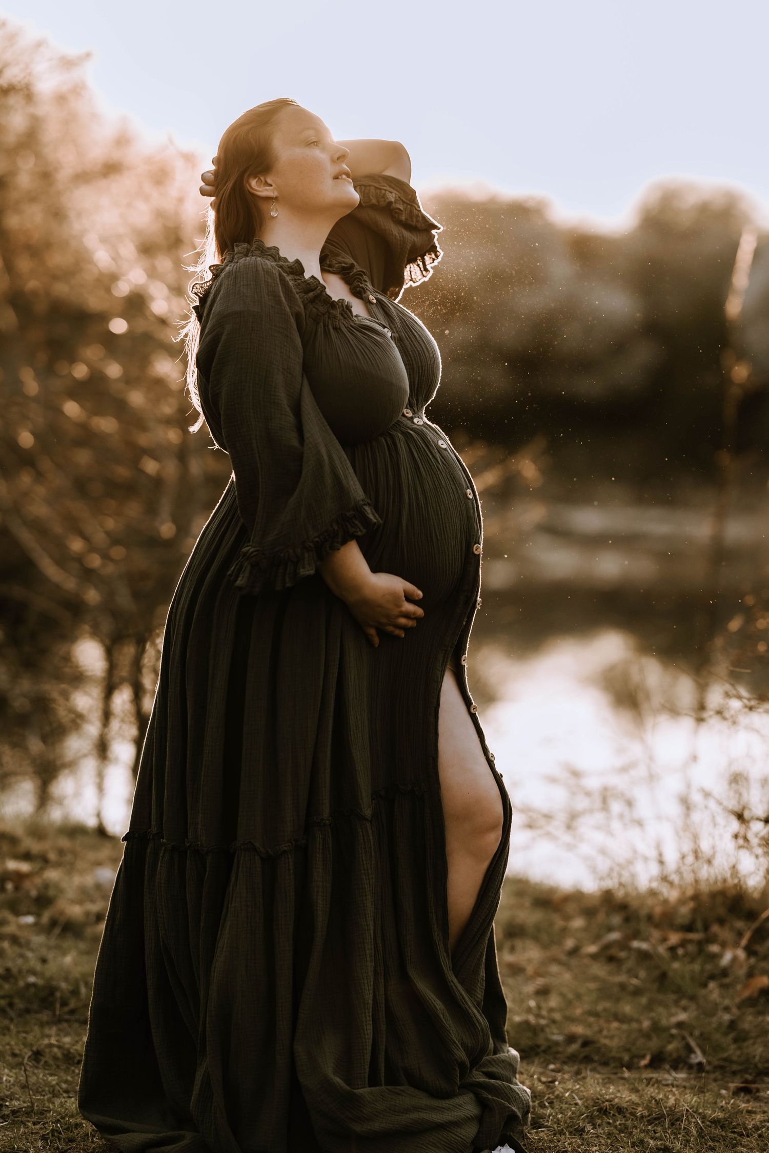 Silhouetted maternity figure in flowing dress against golden sunset by water.