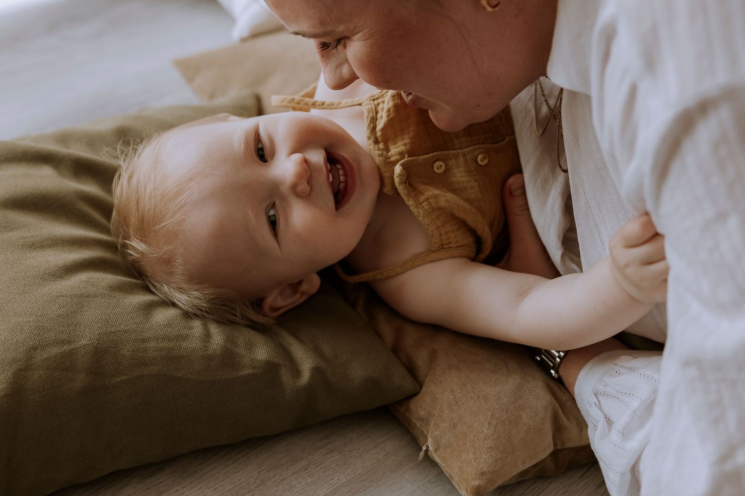 Baby in yellow outfit lies on pillow while parent leans in close.