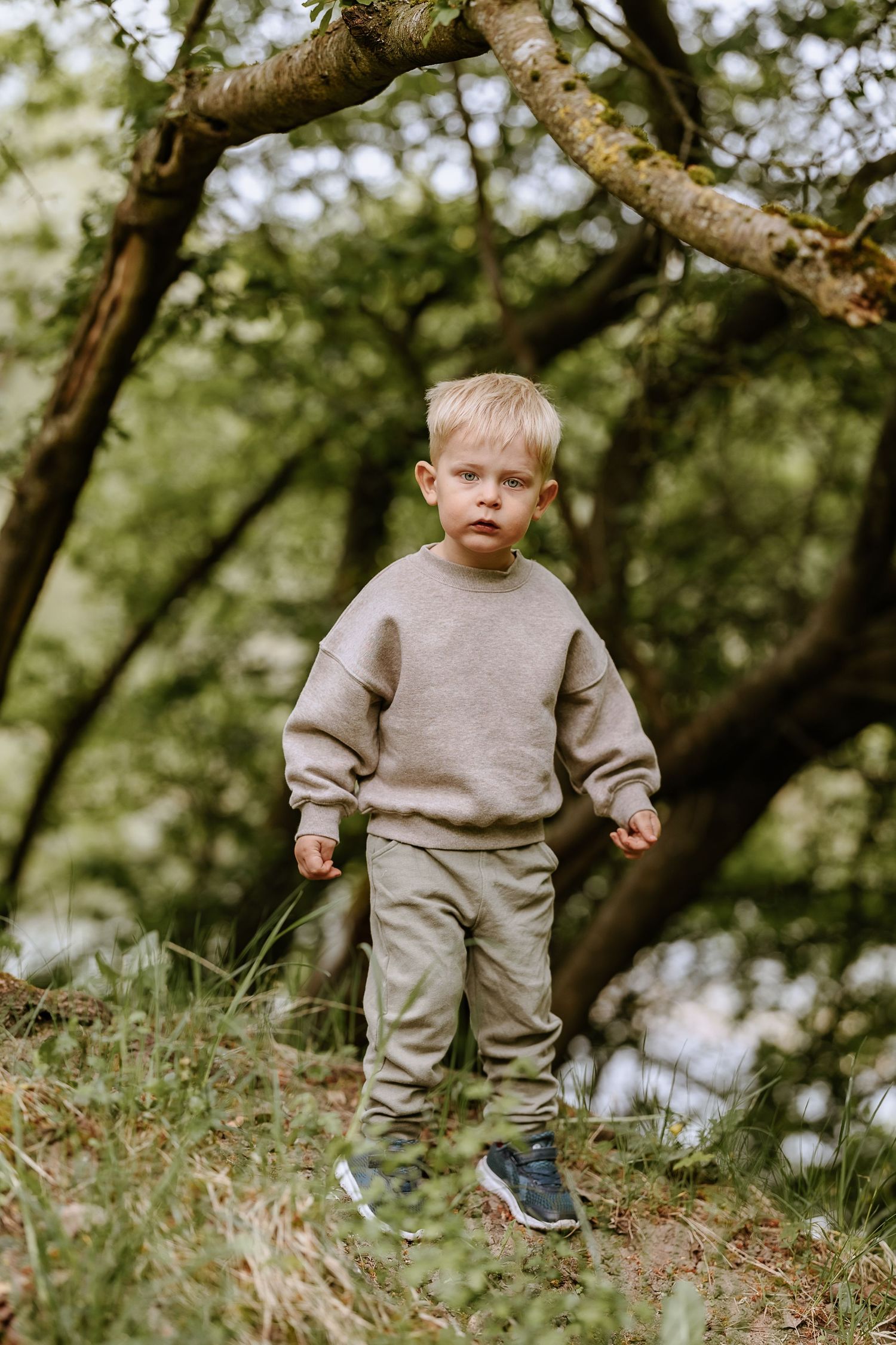 A small child stands alone on a grassy slope wearing a beige sweatsuit and blue sneakers.