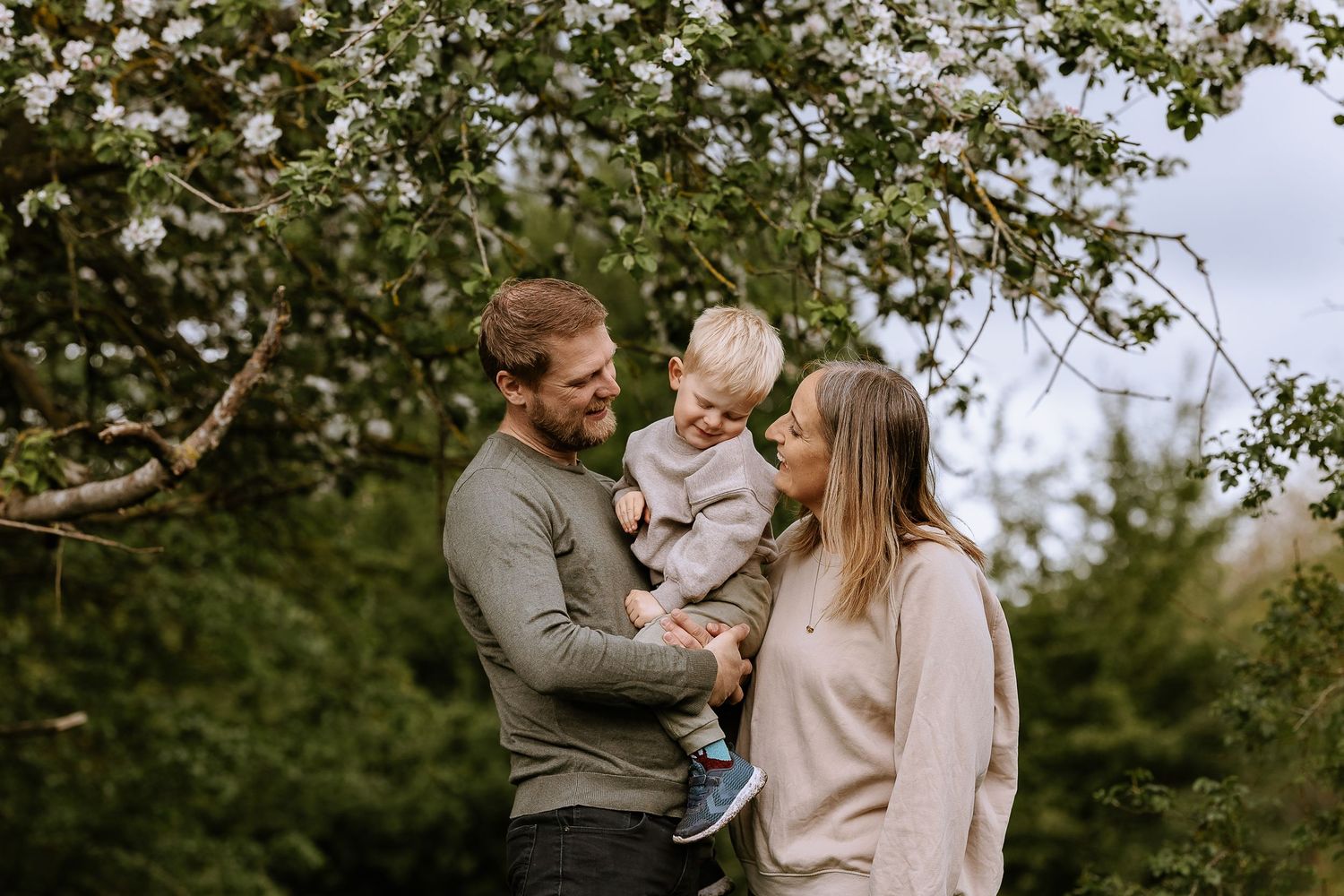 A couple shares tender moments with their baby while standing in a lush green outdoor setting during autumn.