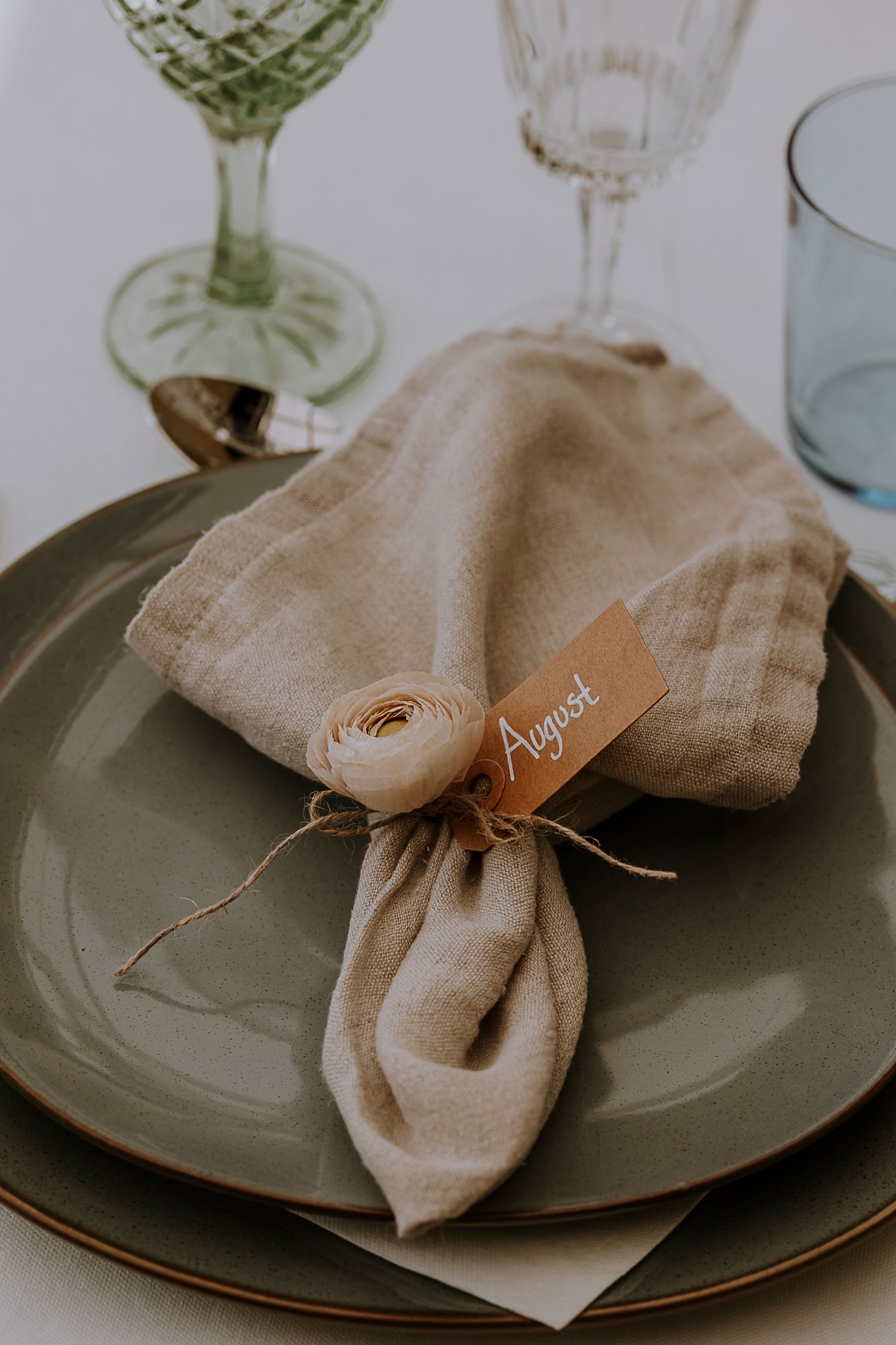 Place setting with beige linen napkin and name card decorated with dried flower on gray plate.