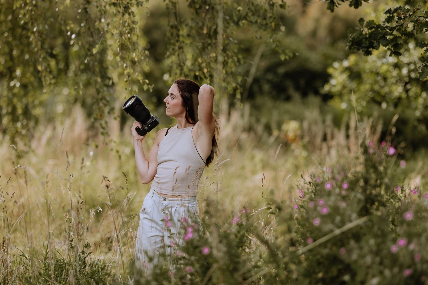 A photographer in a white dress stands in a sunlit meadow with tall grass and wildflowers while holding a camera.
