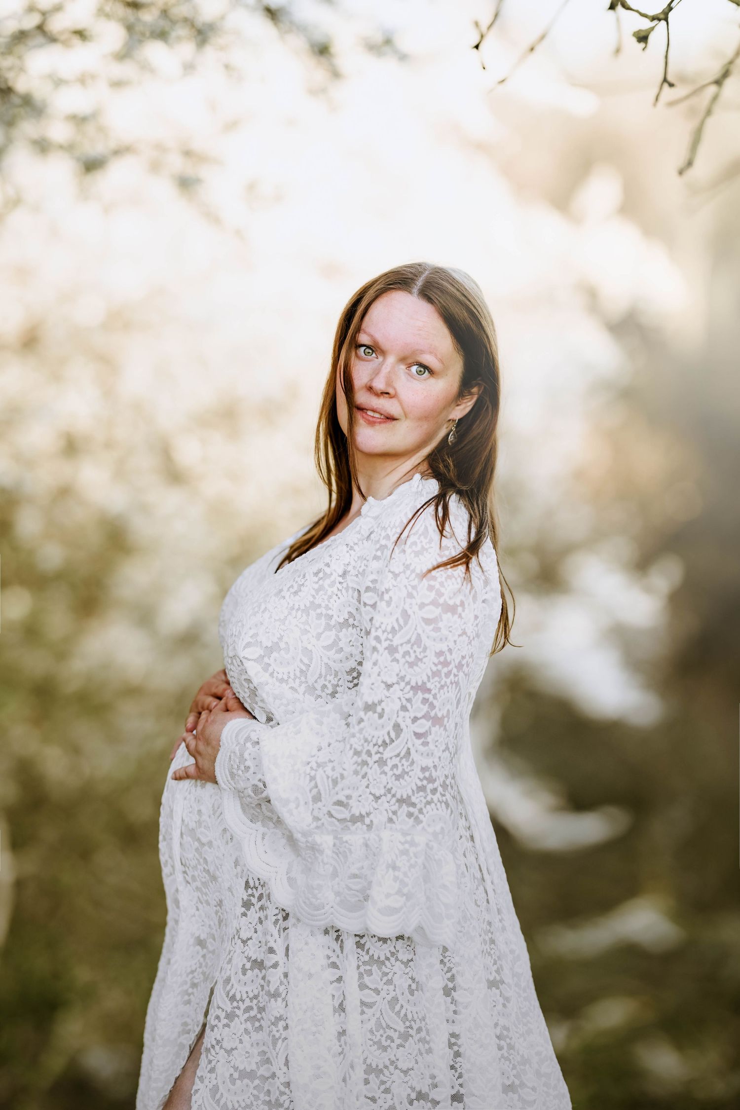 Expecting mother in white lace dress poses in front of blooming spring trees at sunset.