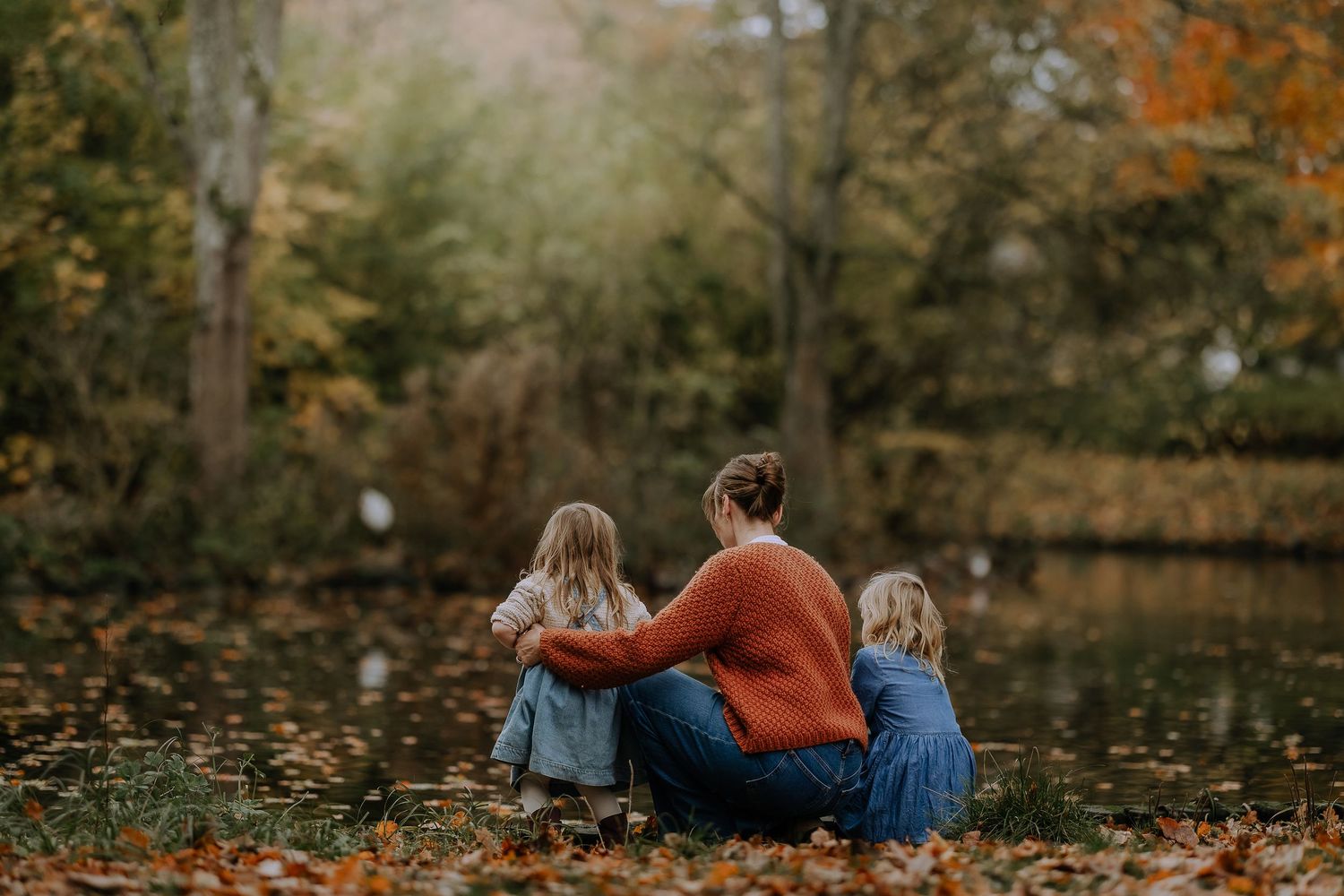 Two people sit close together on fallen autumn leaves in a forest setting during fall season.
