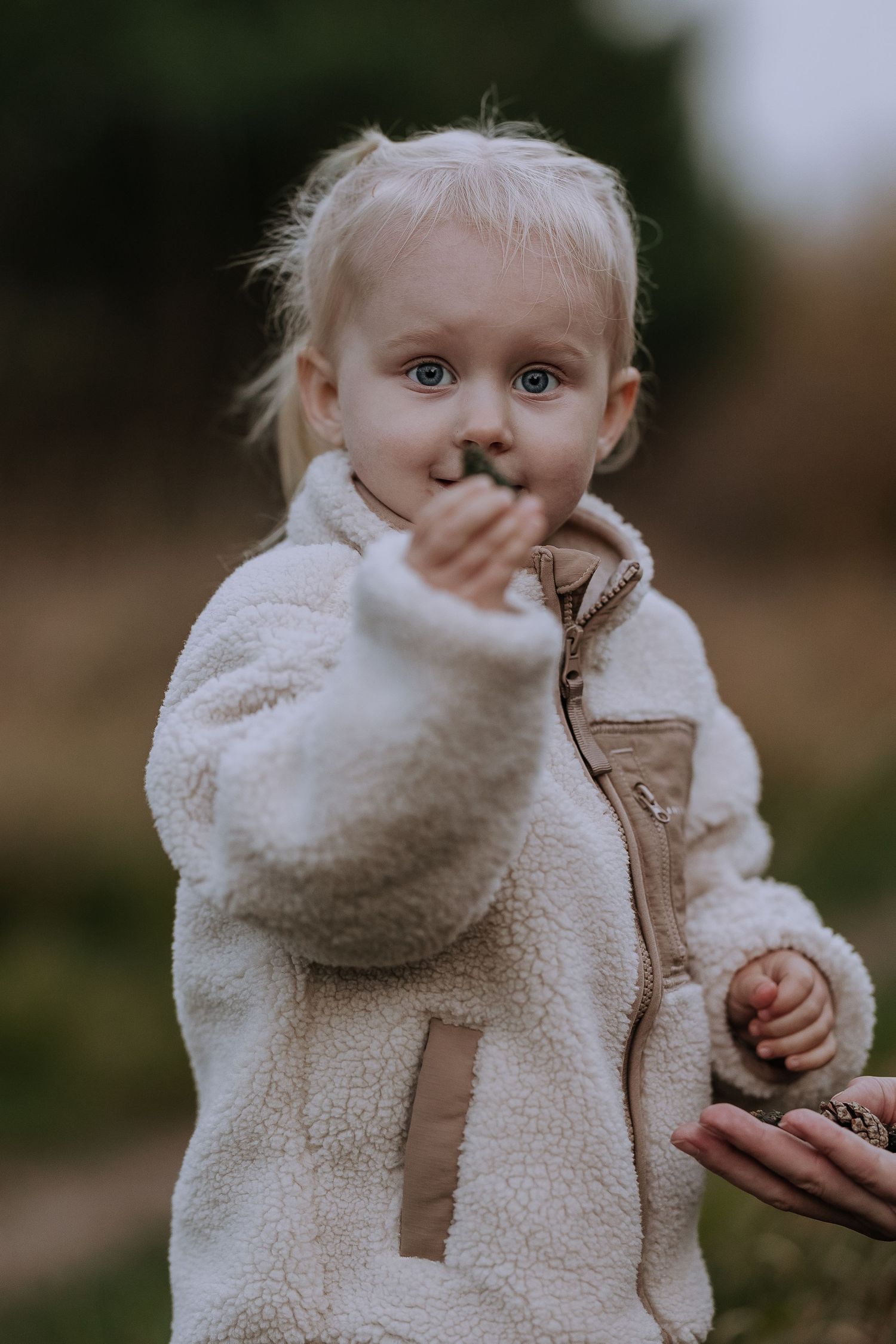Young child in a cozy cream sherpa jacket makes a playful gesture outdoors against a blurred natural background.