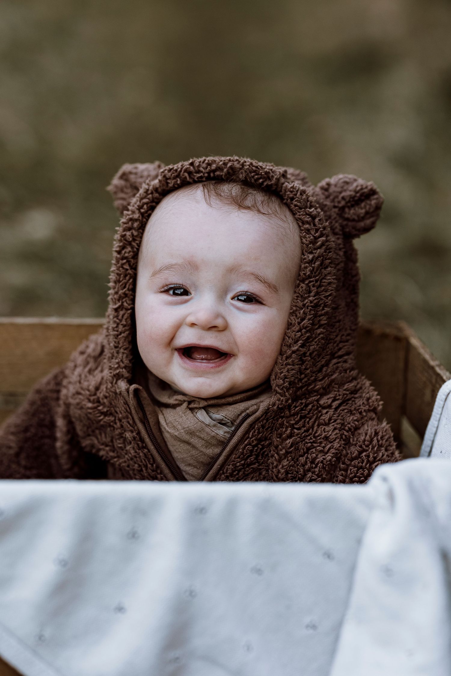 A smiling baby wearing a cozy brown teddy bear hooded outfit lying on white bedding.
