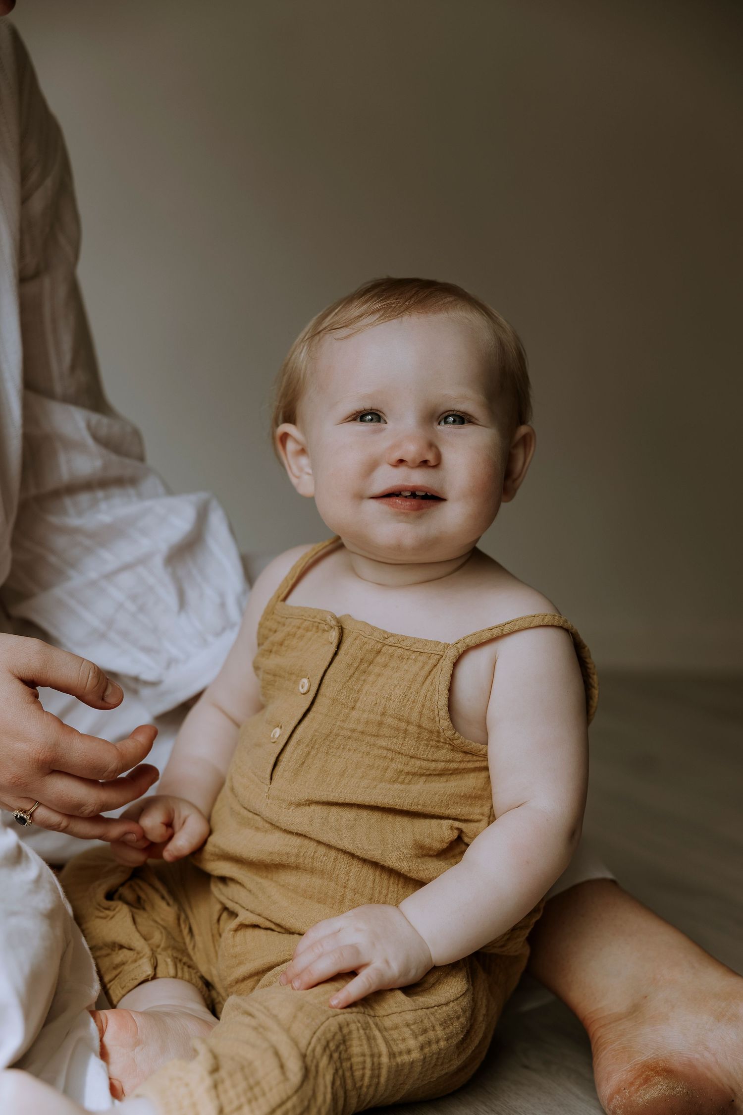 A happy baby in a tan romper sits on a bed while being supported by an adult's hand.
