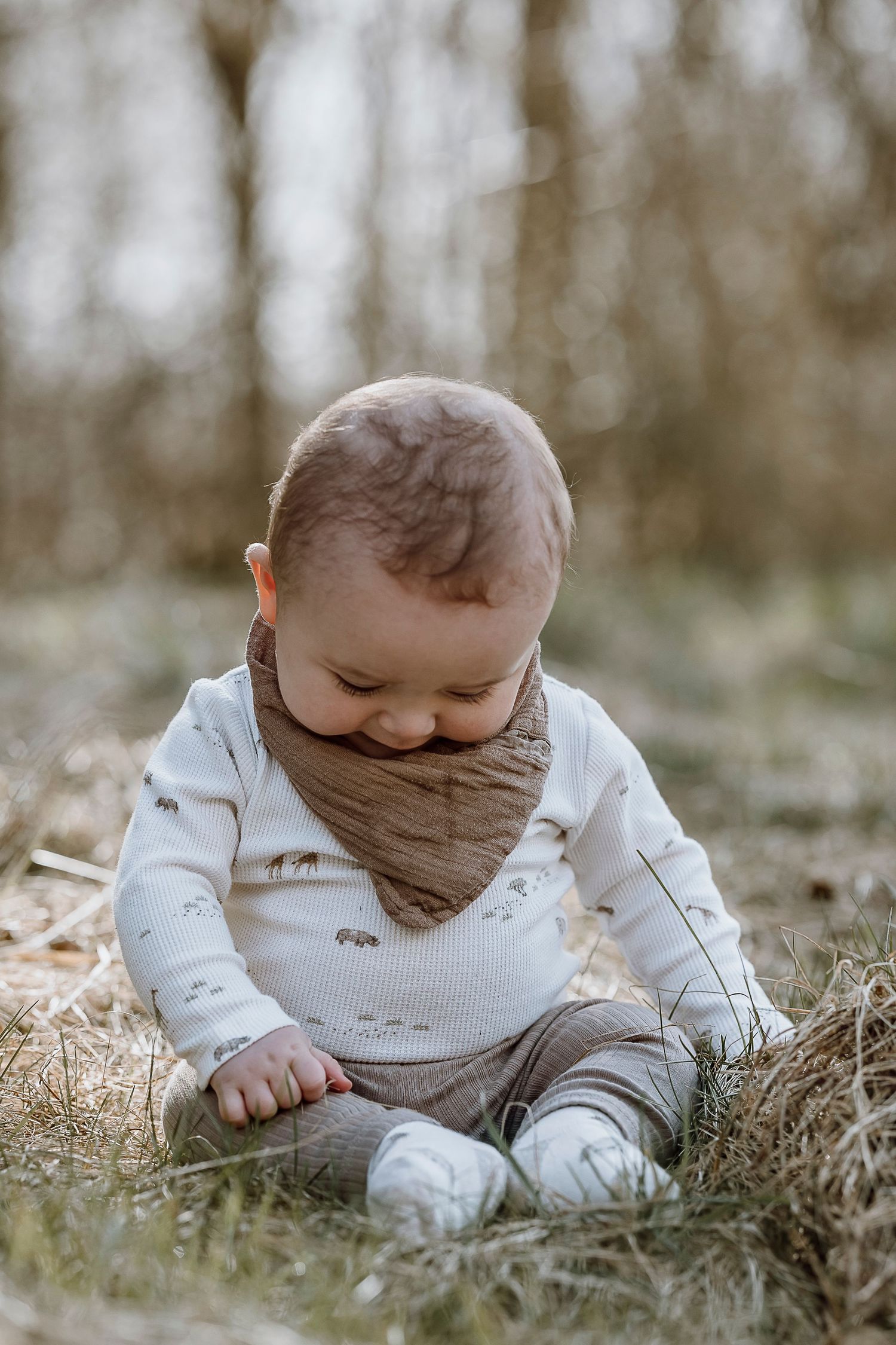 Baby in white sweater and brown bandana bib sits in grass looking down at ground during outdoor photo session.