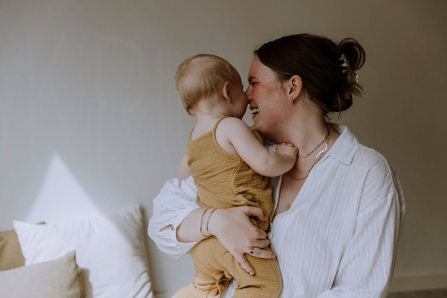 Parent in white shirt embraces baby wearing mustard yellow romper while sitting on bed with pillows.