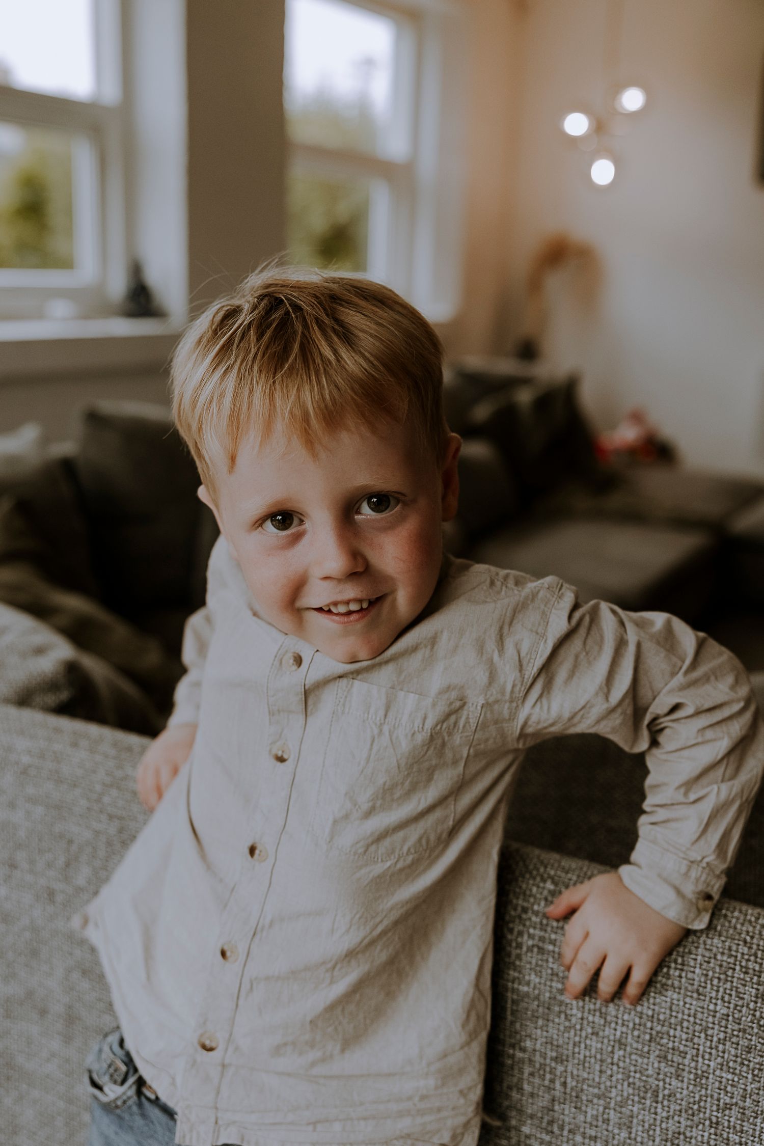 Young child in white button-up shirt smiles while leaning on grey couch.