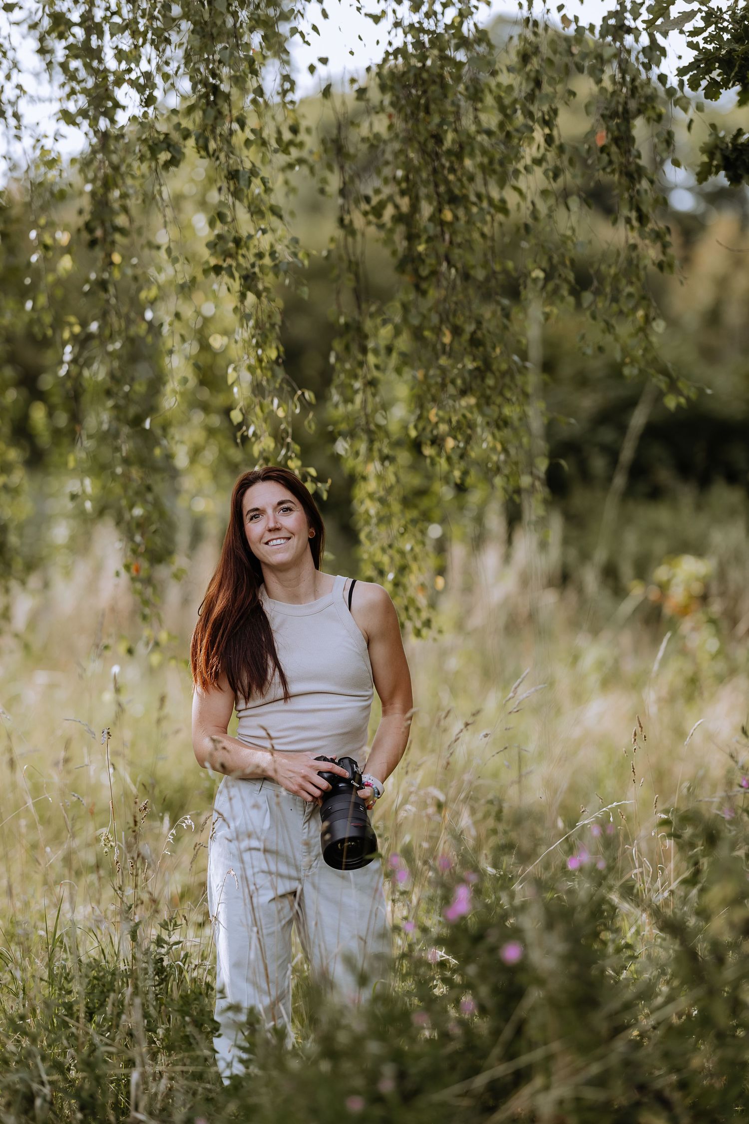 A photographer in a light-colored outfit sits among tall grass and wildflowers in a natural outdoor setting.