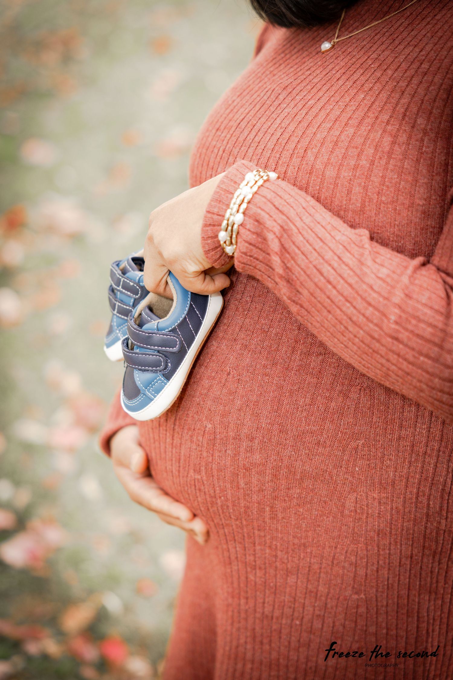 Close-up of baby blue shoes held against a rust-colored maternity sweater with fall foliage in background.