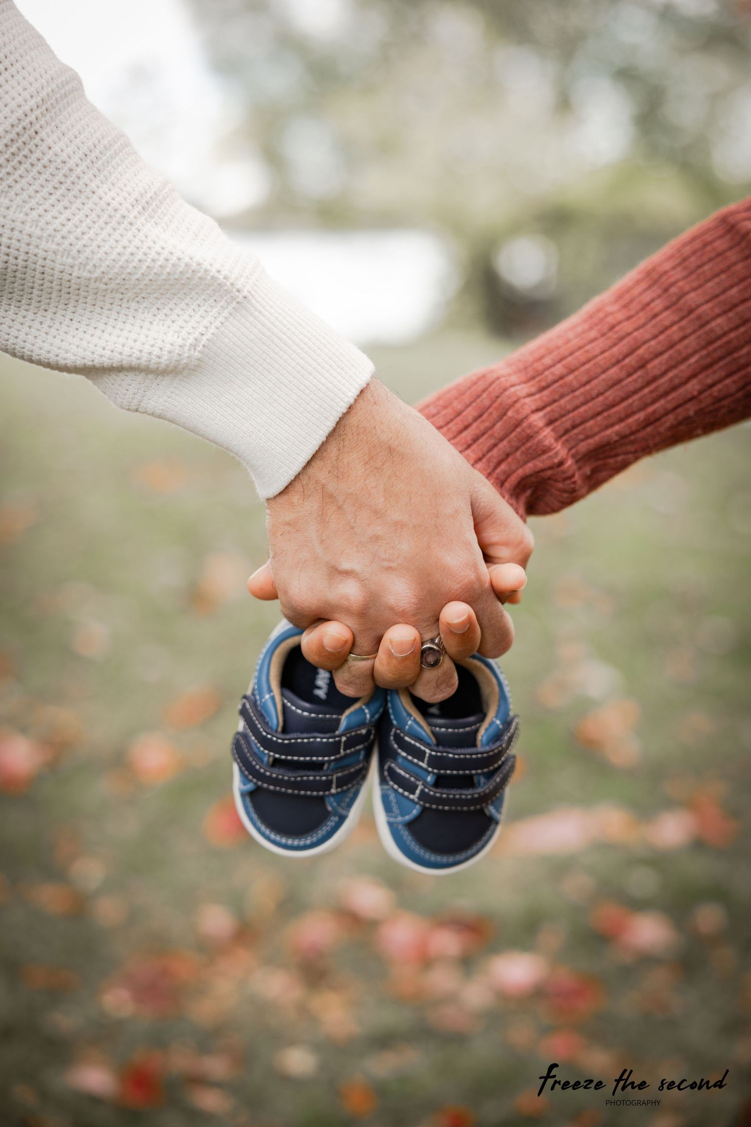 Hands holding tiny blue baby shoes against autumn background with fallen leaves.