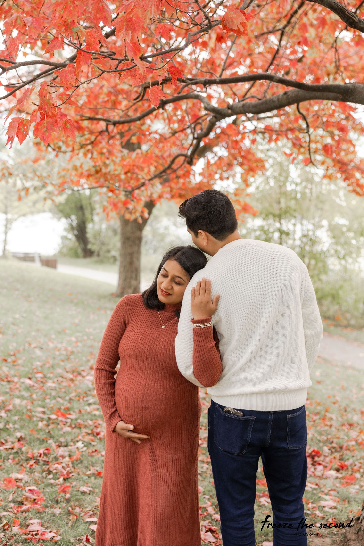 Autumn maternity photo session showing couple in coordinating outfits under red maple trees.