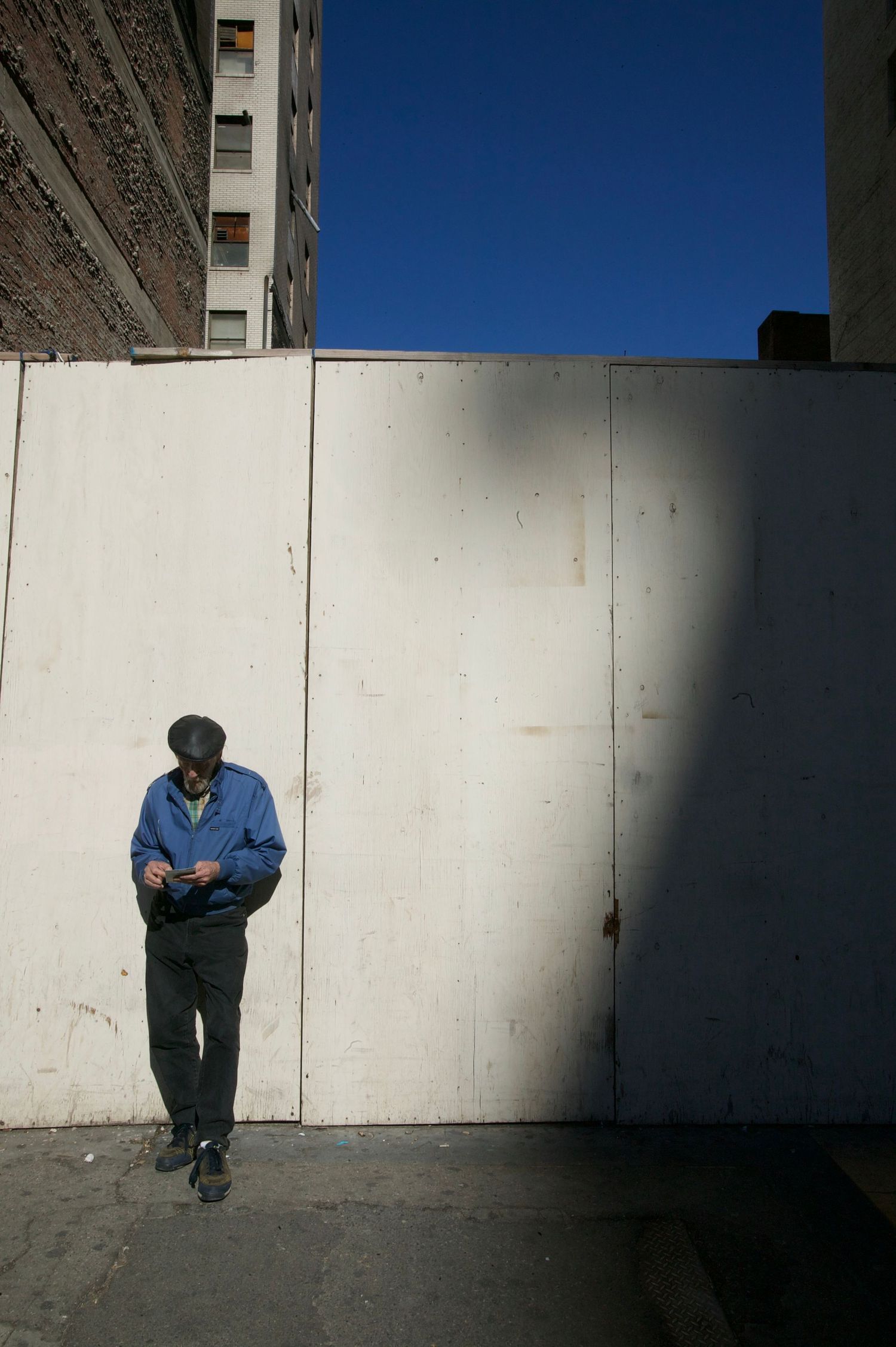 Person in blue jacket standing alone against white wall in harsh sunlight.