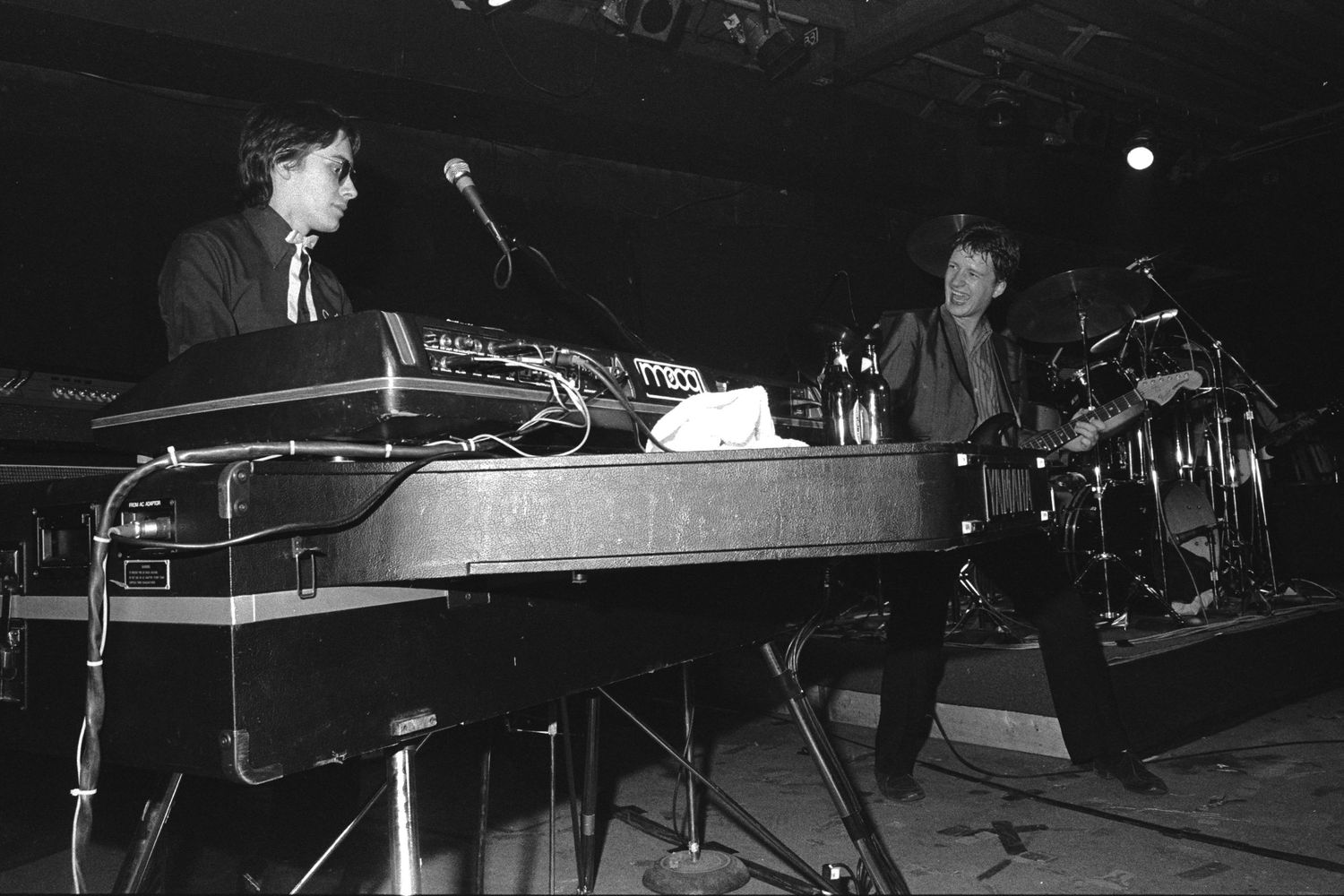 Musicians perform on a dimly lit stage with guitars and microphones in vintage black and white photography.