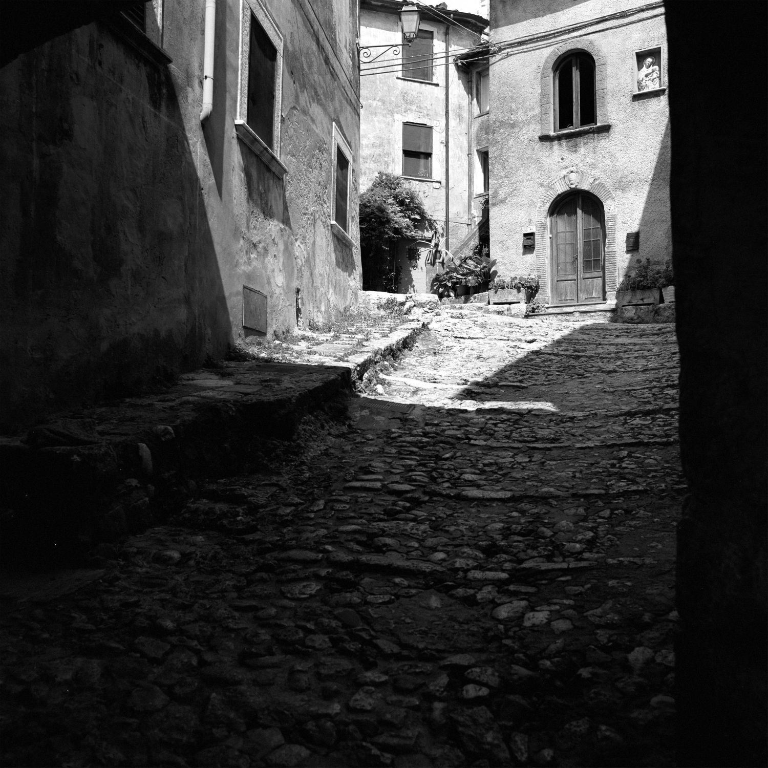 Shadowy cobblestone alley between old stone buildings in dramatic black and white.