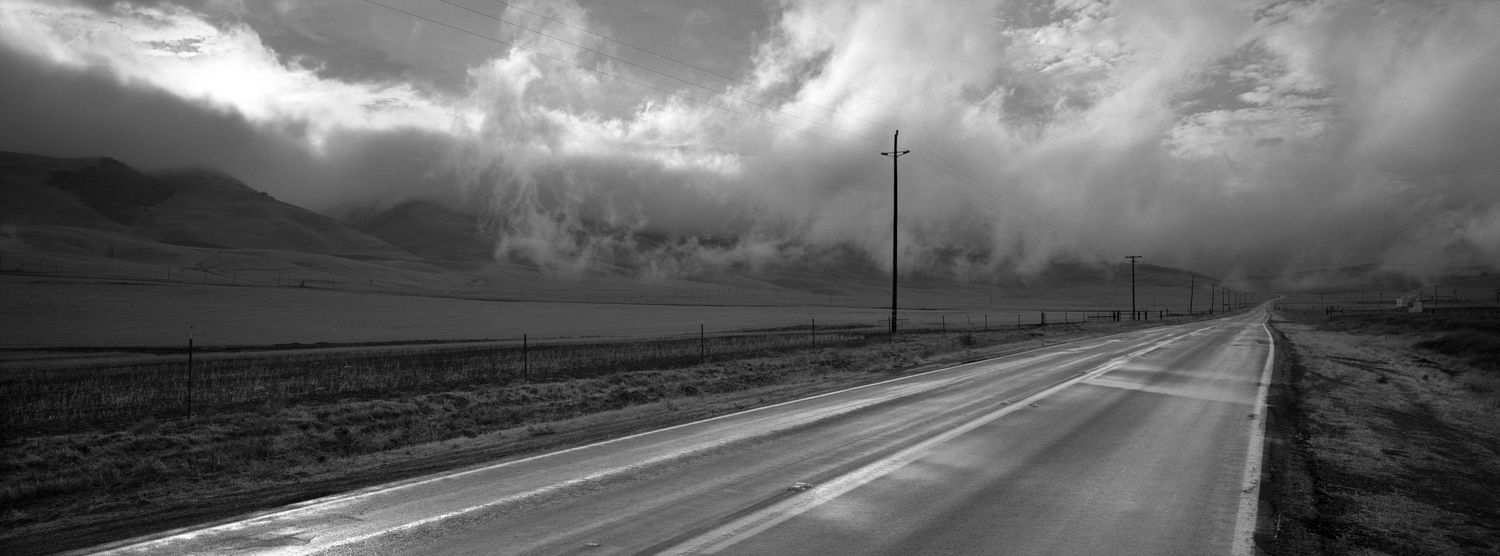 A long empty highway stretches into the distance under dramatic storm clouds and mountain peaks.