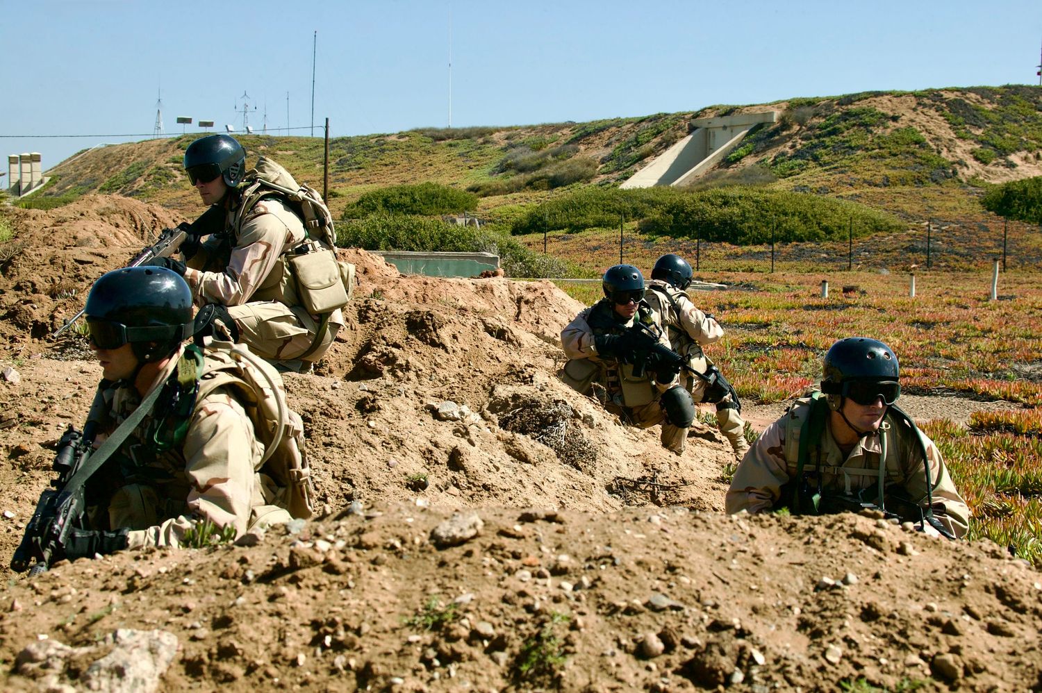 Military soldiers in desert camouflage gear take defensive positions behind rocky terrain during a training exercise.