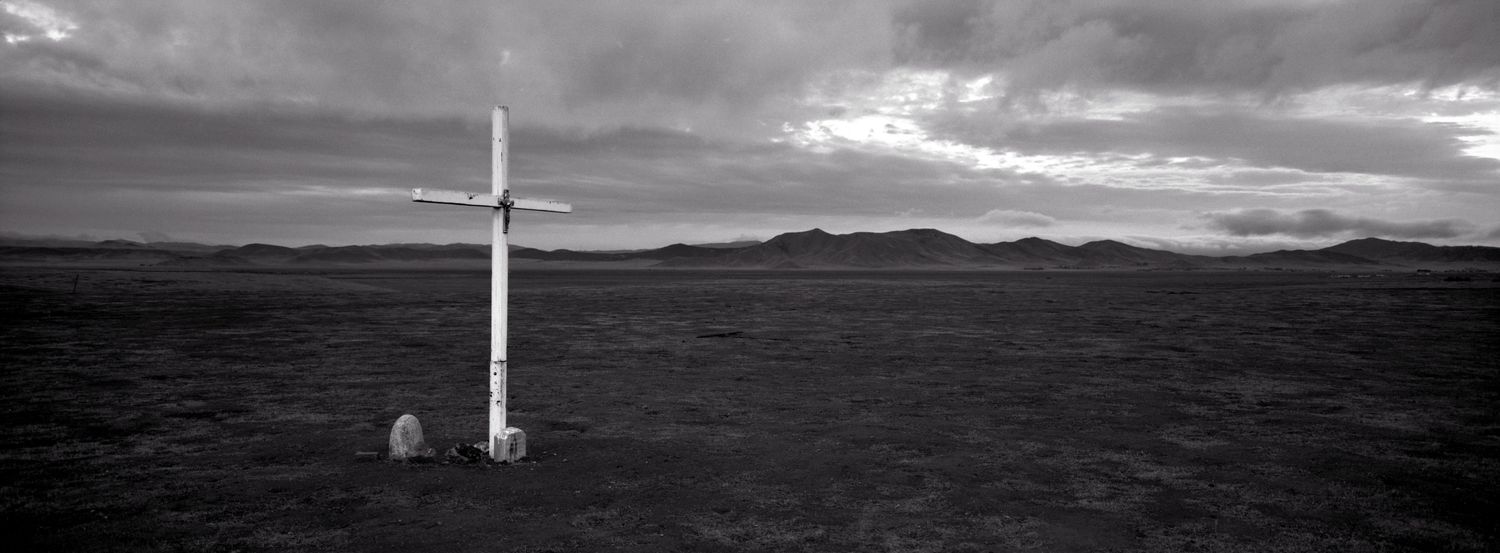 A panoramic black and white landscape shows a dramatic cloudy sky over rolling hills with a single cross monument.