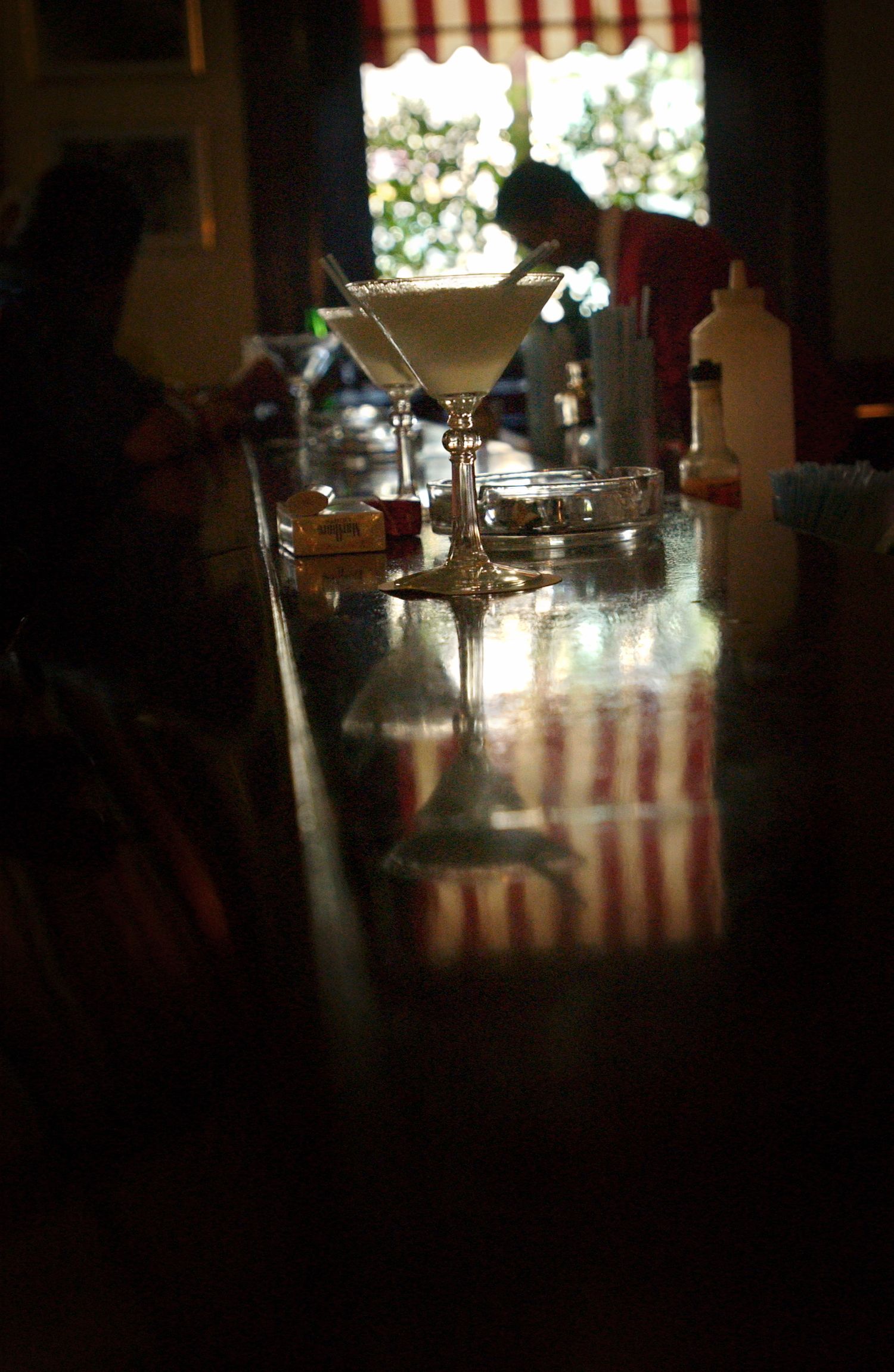 Dark atmospheric bar interior with glasses and bottles silhouetted against window light.